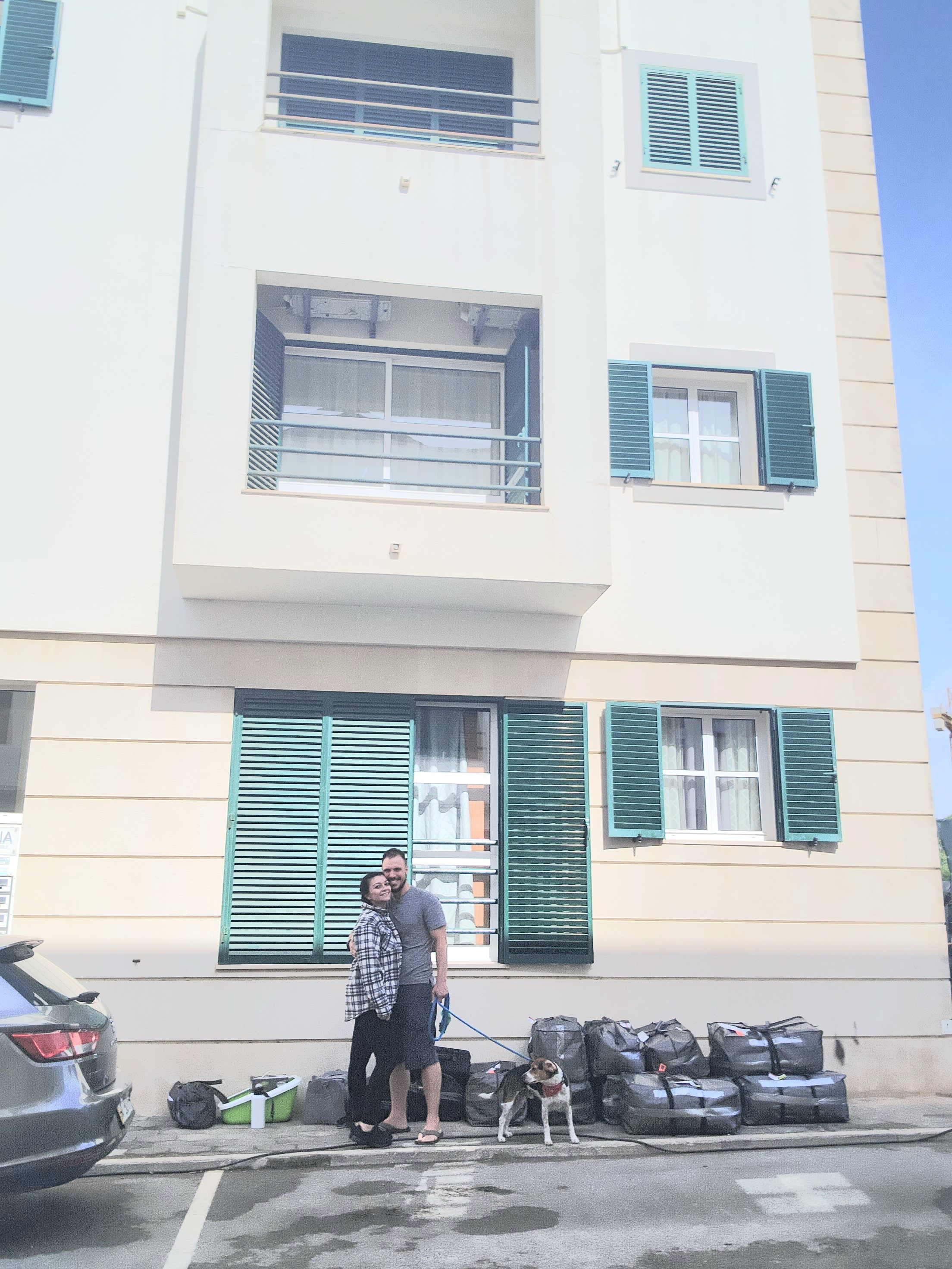 A couple stands happily in front of a beige building with green shutters, accompanied by a dog on a leash. Several packed bags are lined up on the sidewalk next to them. A car is parked nearby, and the scene is set under a clear blue sky.
