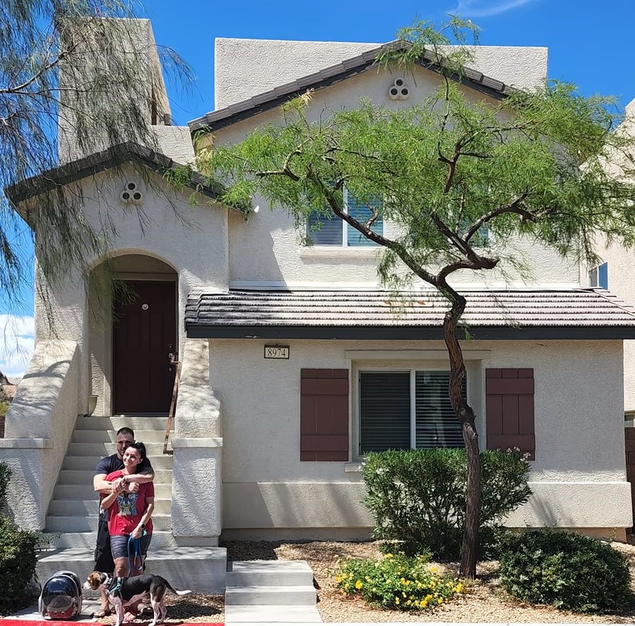 A couple stands happily in front of a beige, two-story house with maroon shutters and a small tree in the yard. They are accompanied by a dog and a pet carrier on the sidewalk. The house features a red front door and is surrounded by neatly trimmed bushes under a clear blue sky.