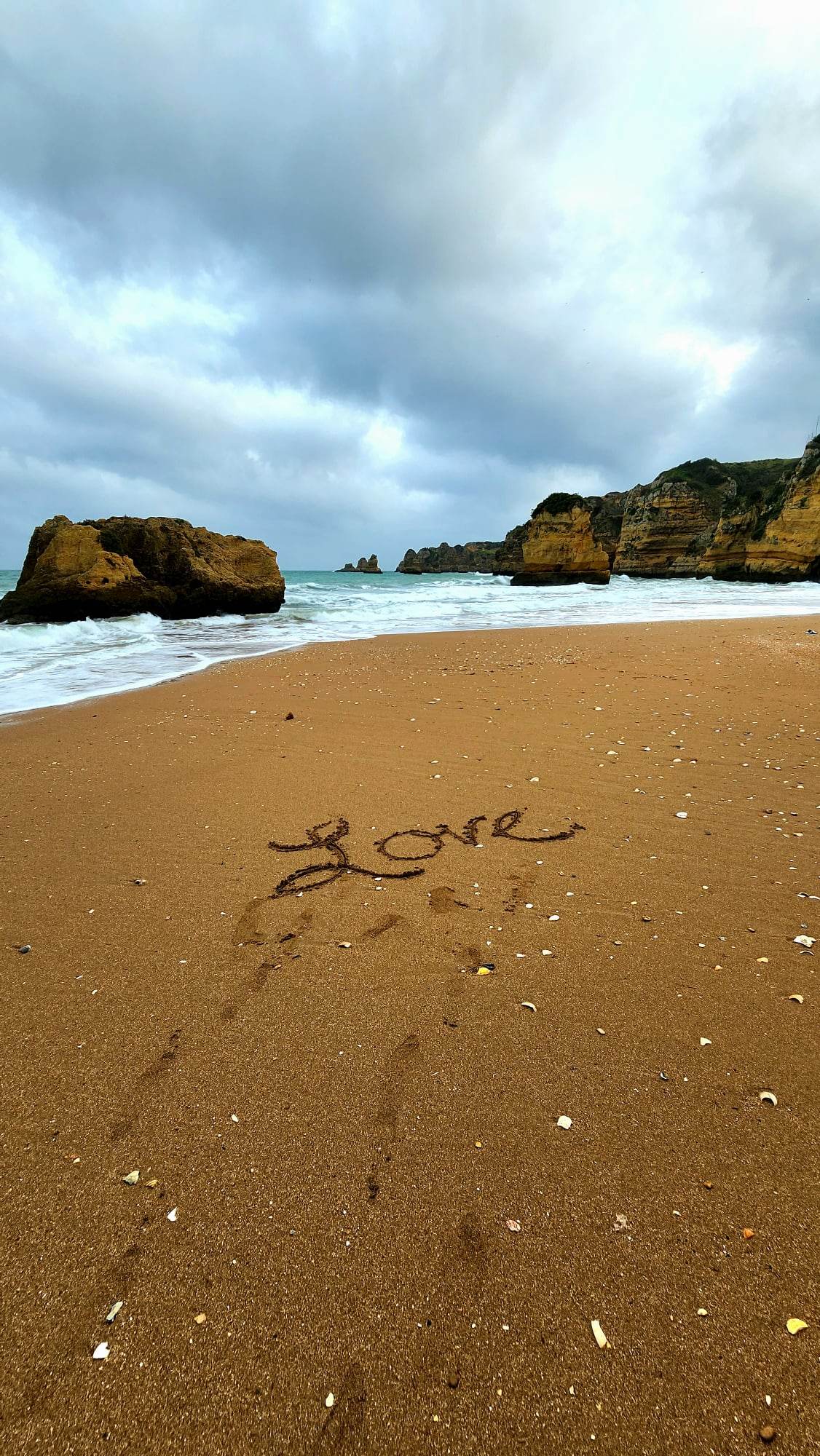 The word "Love" is written in the sand on a deserted beach, with scattered seashells and footprints leading towards the shoreline, where waves gently lap against large rocky formations under a cloudy sky.