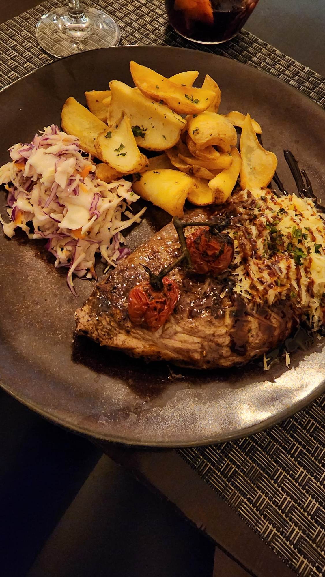 A delicious plate featuring a juicy steak topped with roasted cherry tomatoes and grated cheese, accompanied by crispy potato wedges and a side of colorful coleslaw. The dish is elegantly presented on a dark plate, with a glass of red drink in the background.