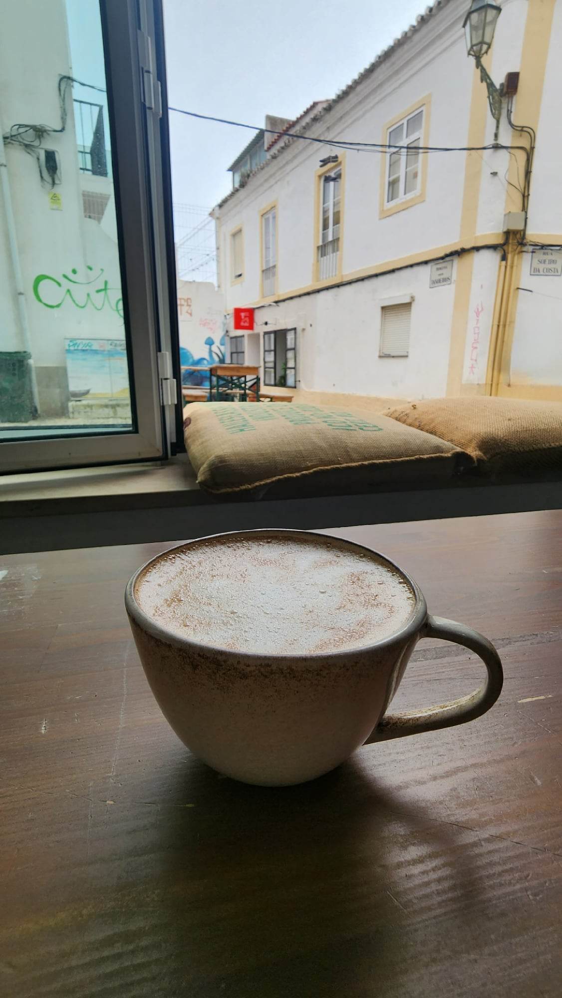 A frothy cappuccino in a rustic ceramic mug sits on a wooden table by an open window, revealing a view of a quaint street with white buildings and graffiti.