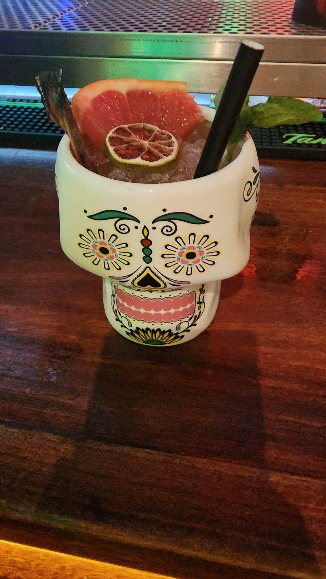 A decorative skull-shaped mug sits on a wooden bar, filled with a cocktail garnished with a grapefruit slice and a dried lime. A black straw and a sprig of mint add to the vibrant presentation.
