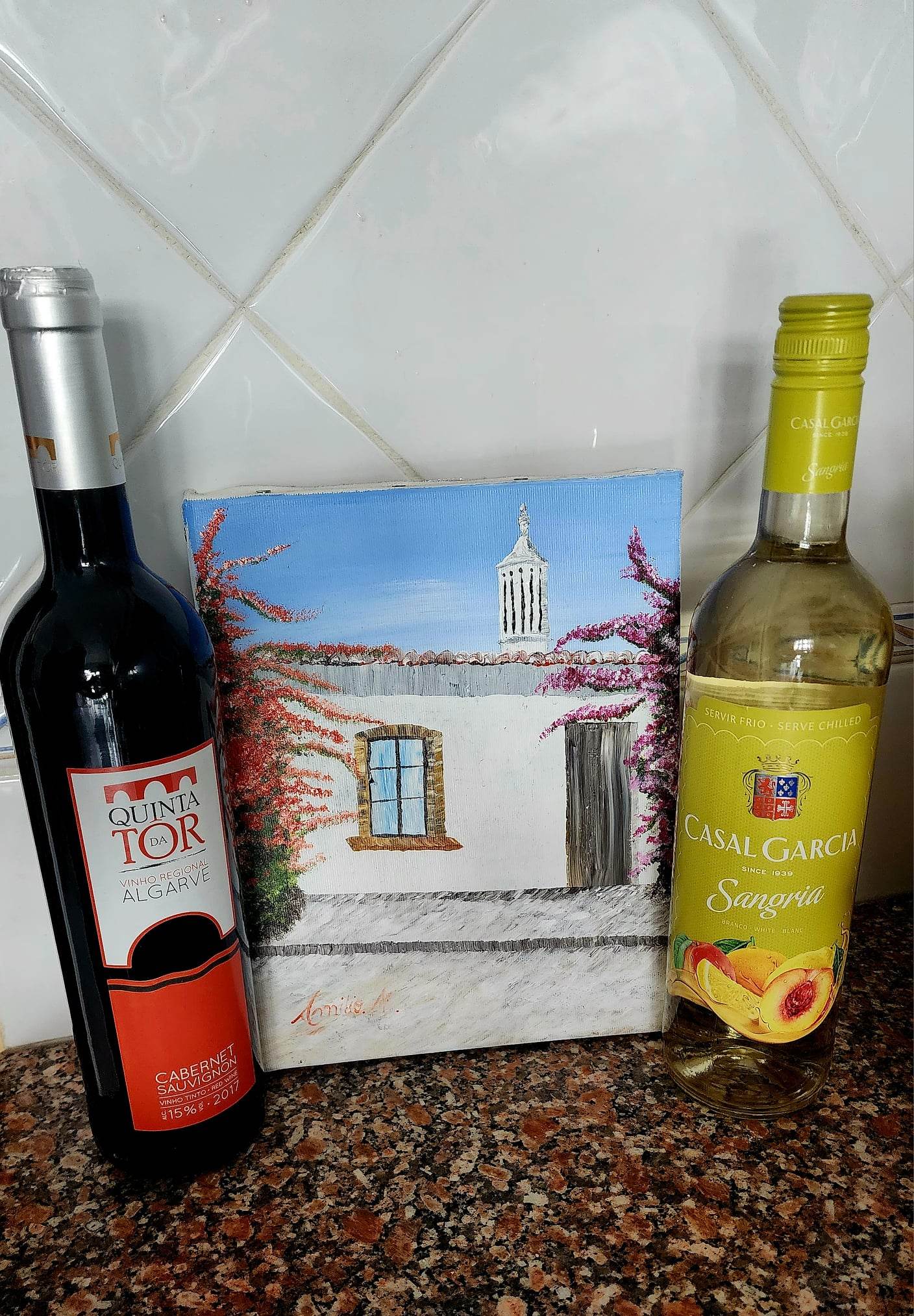 Two wine bottles flank a small painting on a tiled countertop. On the left, a bottle of Quinta da Tôr Cabernet Sauvignon stands with a red label, while on the right, a bottle of Casal Garcia Sangria with a yellow label. The painting depicts a charming white house with a blue sky and flowering vines.