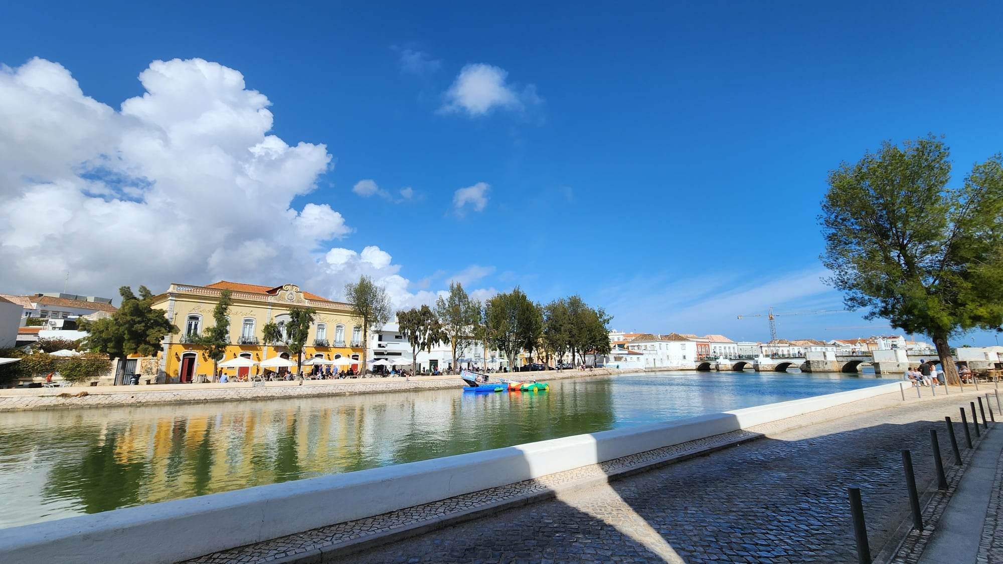 A picturesque riverside scene features a charming yellow building with outdoor seating and umbrellas, reflecting in the calm water. Trees line the riverbank, and a stone bridge arches over the river under a vibrant blue sky with fluffy clouds.
