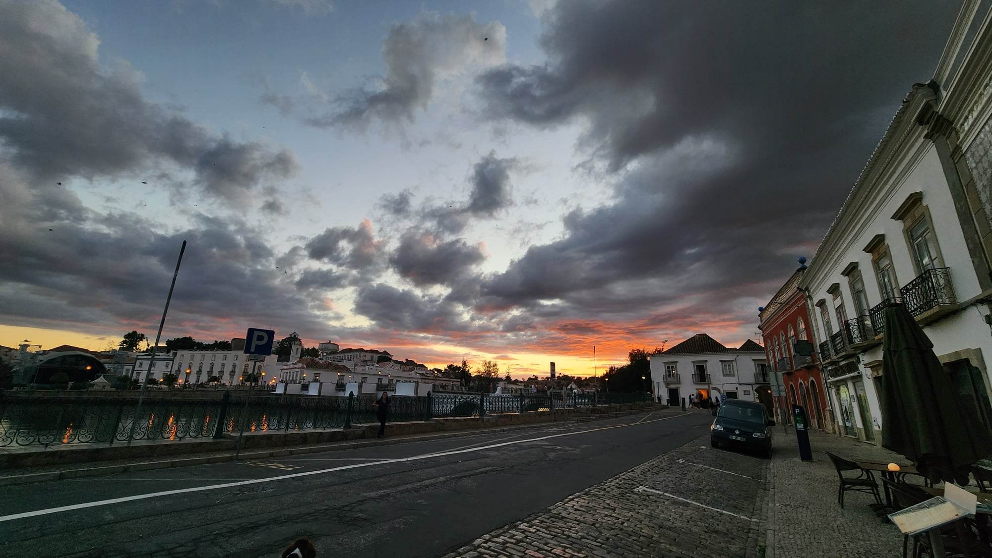 A picturesque street scene at sunset, with a dramatic sky filled with dark, scattered clouds and a vibrant orange horizon. On the right, charming buildings with balconies line the cobblestone sidewalk, while a few tables and chairs sit outside. A river runs alongside the street on the left, reflecting the warm glow of the streetlights.