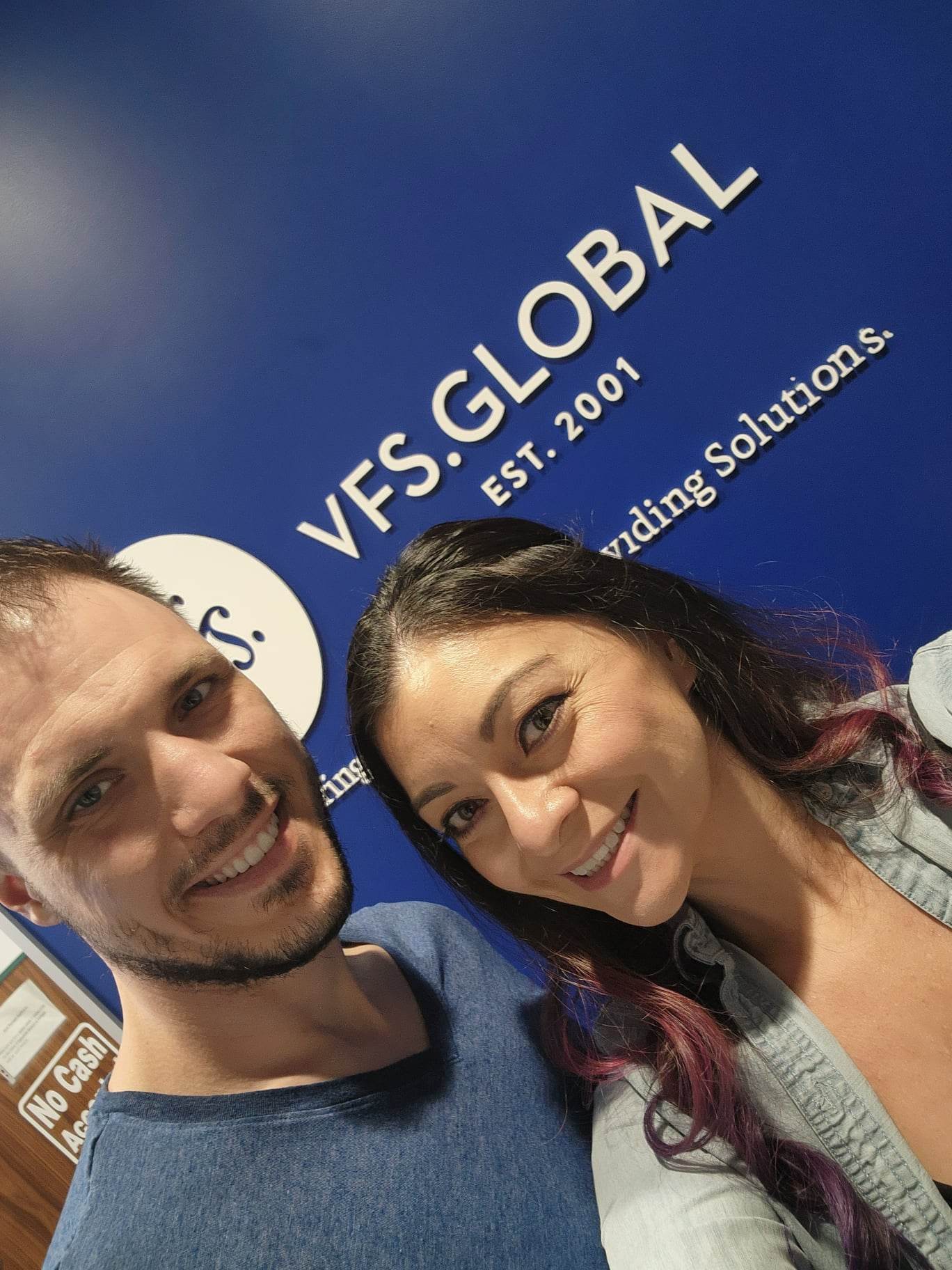 A smiling man and woman take a selfie in front of a blue wall with the "VFS Global" logo and "Est. 2001" text.