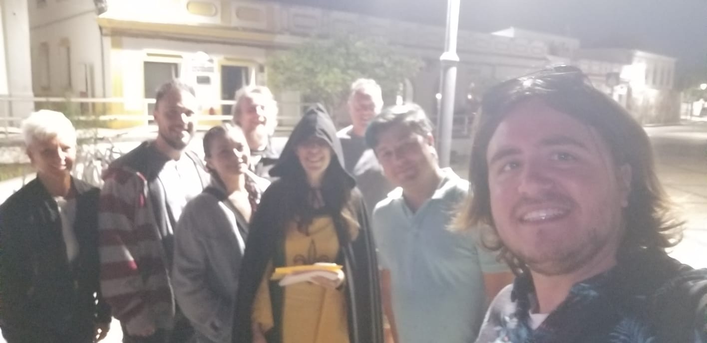 A group of people stands together at night, smiling for a selfie. One person wears a hooded cloak, holding a book. They are gathered under a streetlamp, with a building in the background.