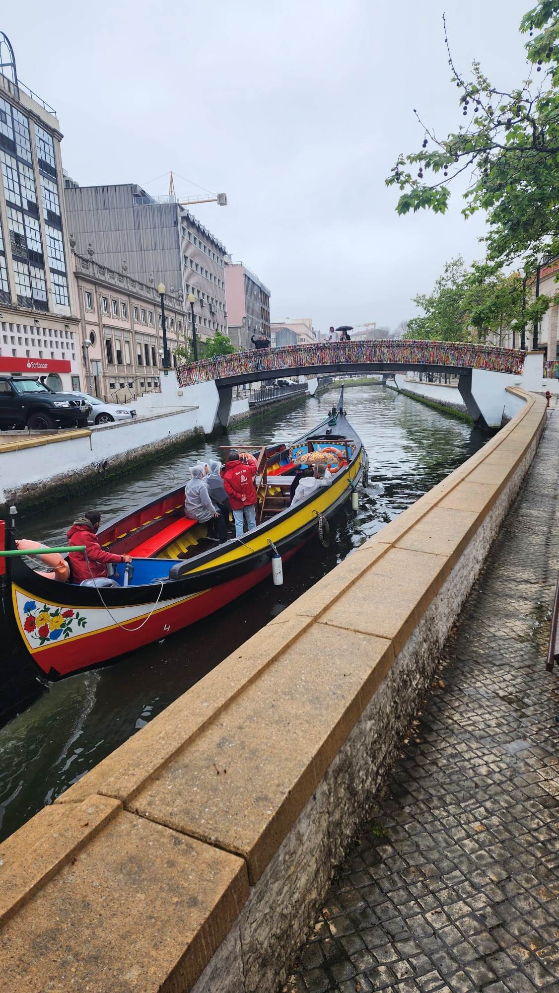 A colorful boat with passengers in raincoats navigates a narrow canal bordered by a cobblestone path. Overhead, a pedestrian bridge adorned with vibrant locks spans the water. Buildings line the canal, and the overcast sky adds a tranquil atmosphere.