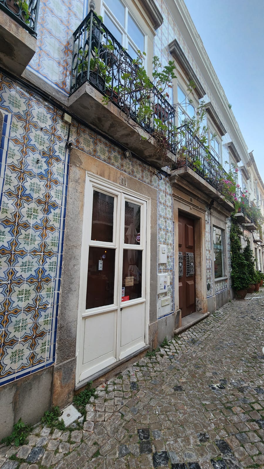 A charming street scene features a building adorned with colorful, intricate tiles and wrought-iron balconies with lush green plants. The cobblestone pathway adds a rustic touch, while a white-framed door and window enhance the quaint atmosphere.