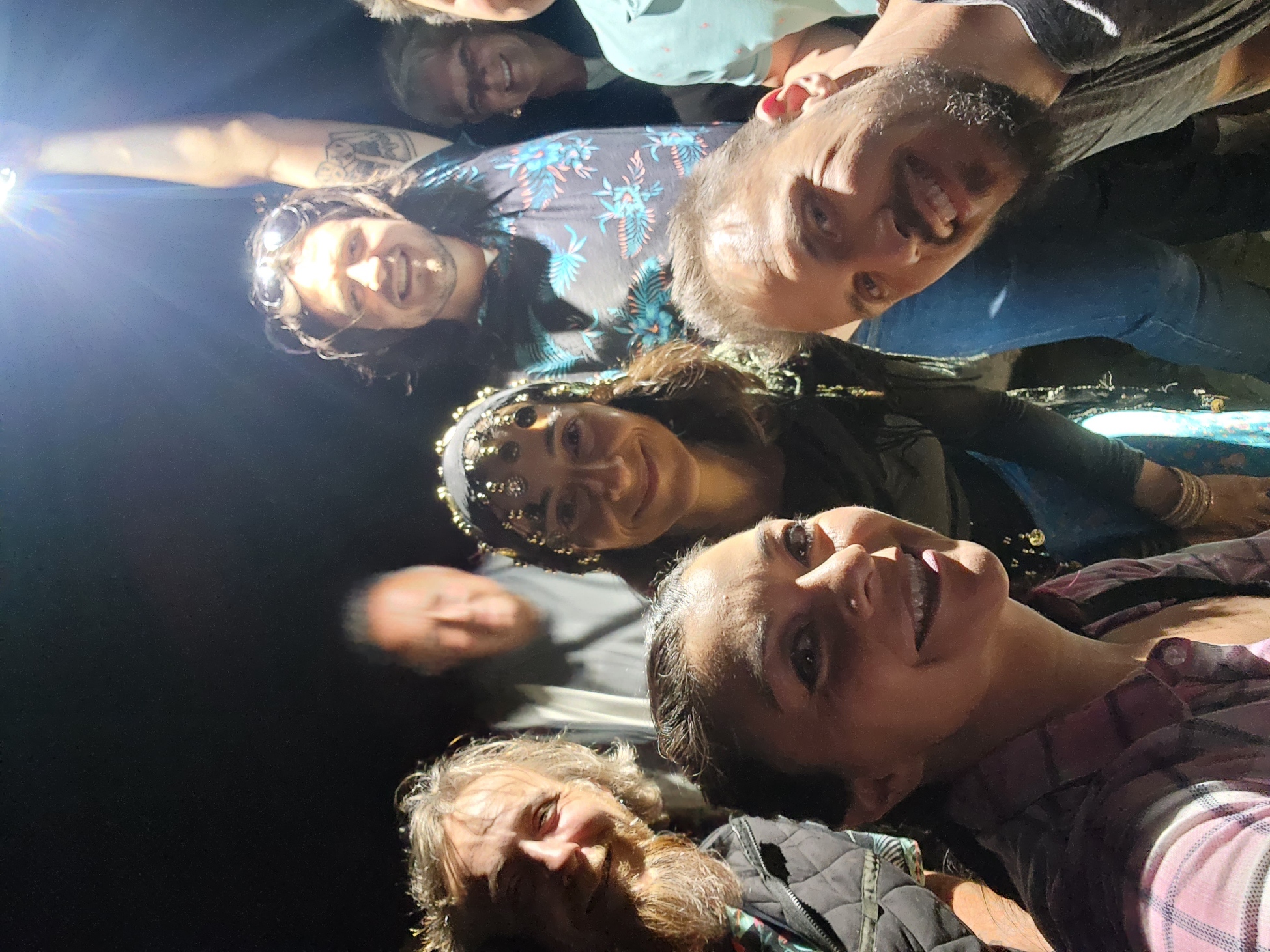 A group of people happily posing for a selfie at night, with one person holding a light above them. The scene includes a woman wearing a headpiece with decorative elements, and others smiling around her. The atmosphere is lively and joyful.