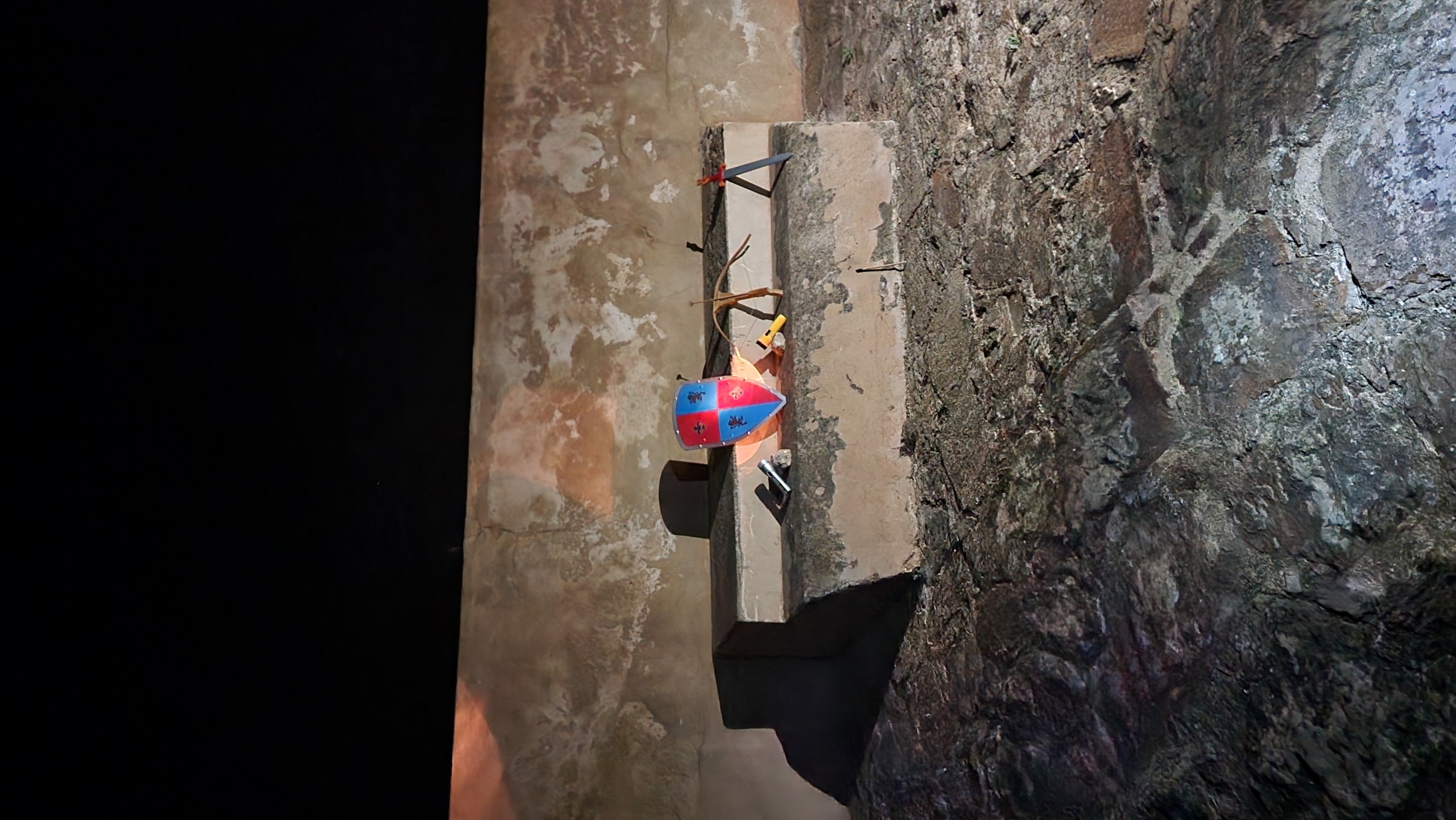 A weathered stone altar stands against a textured wall, adorned with a colorful shield featuring red and blue sections and black symbols. Various tools and objects, including a stick and a small axe, rest on the altar, creating a rustic and historical atmosphere.