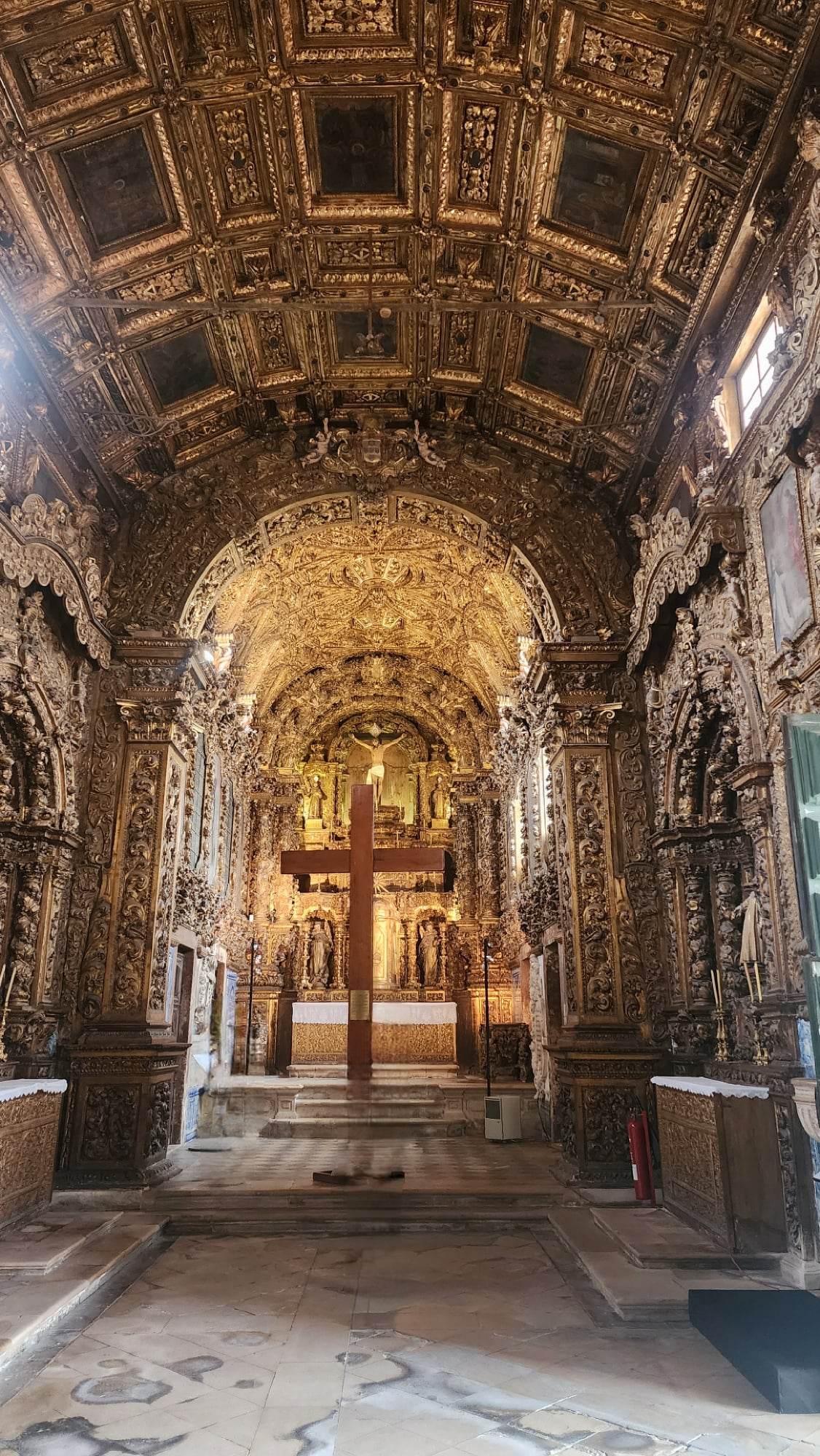 A richly ornate church interior featuring an intricately carved wooden ceiling and walls, all gilded in gold. A large wooden cross stands prominently in the center aisle, leading to an altar adorned with detailed religious sculptures and decorations. Natural light filters in from side windows, highlighting the opulent craftsmanship.