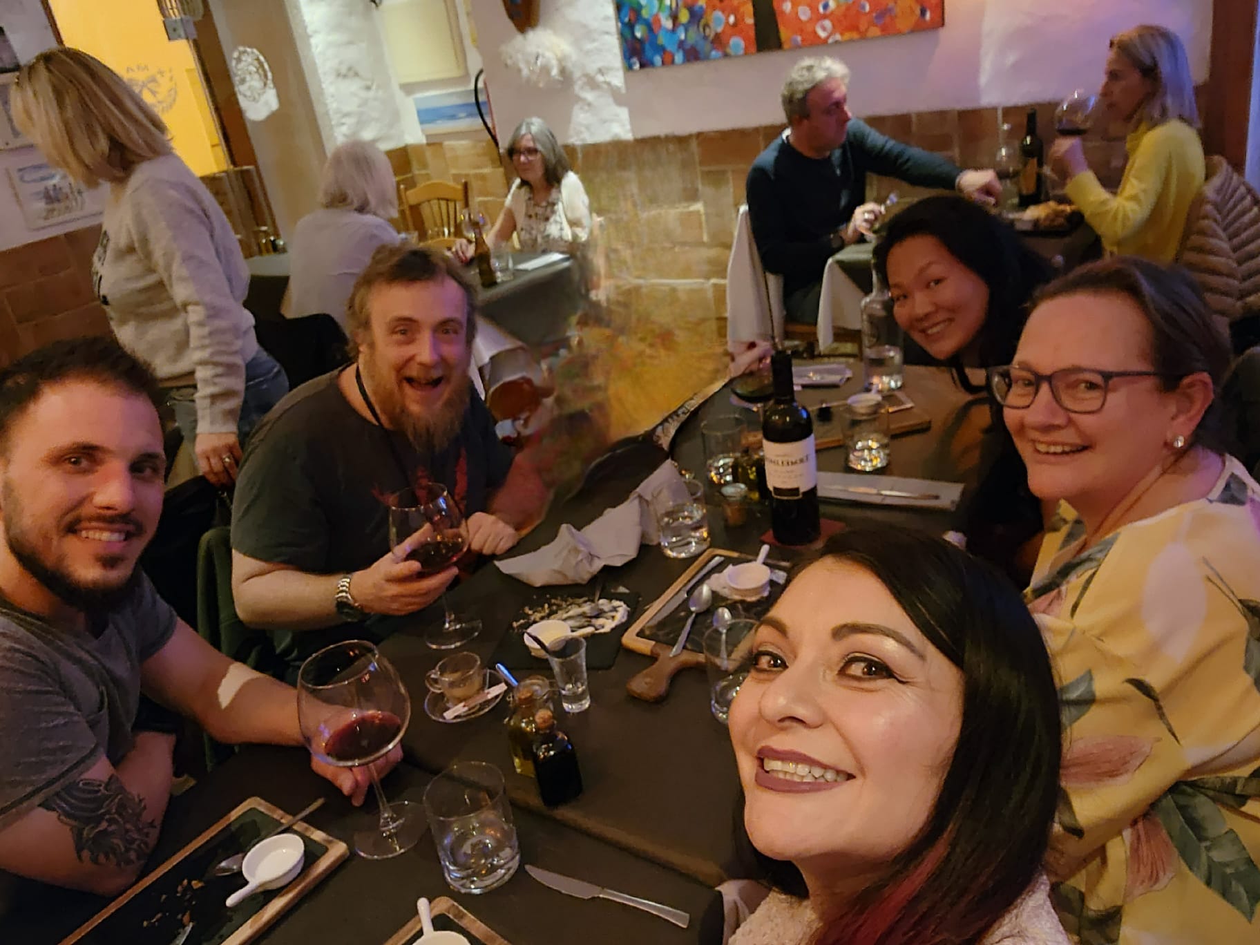 A group of friends is enjoying a meal at a cozy restaurant, smiling and holding glasses of wine. The table is set with plates and a bottle of red wine, and the background features colorful artwork and other diners.