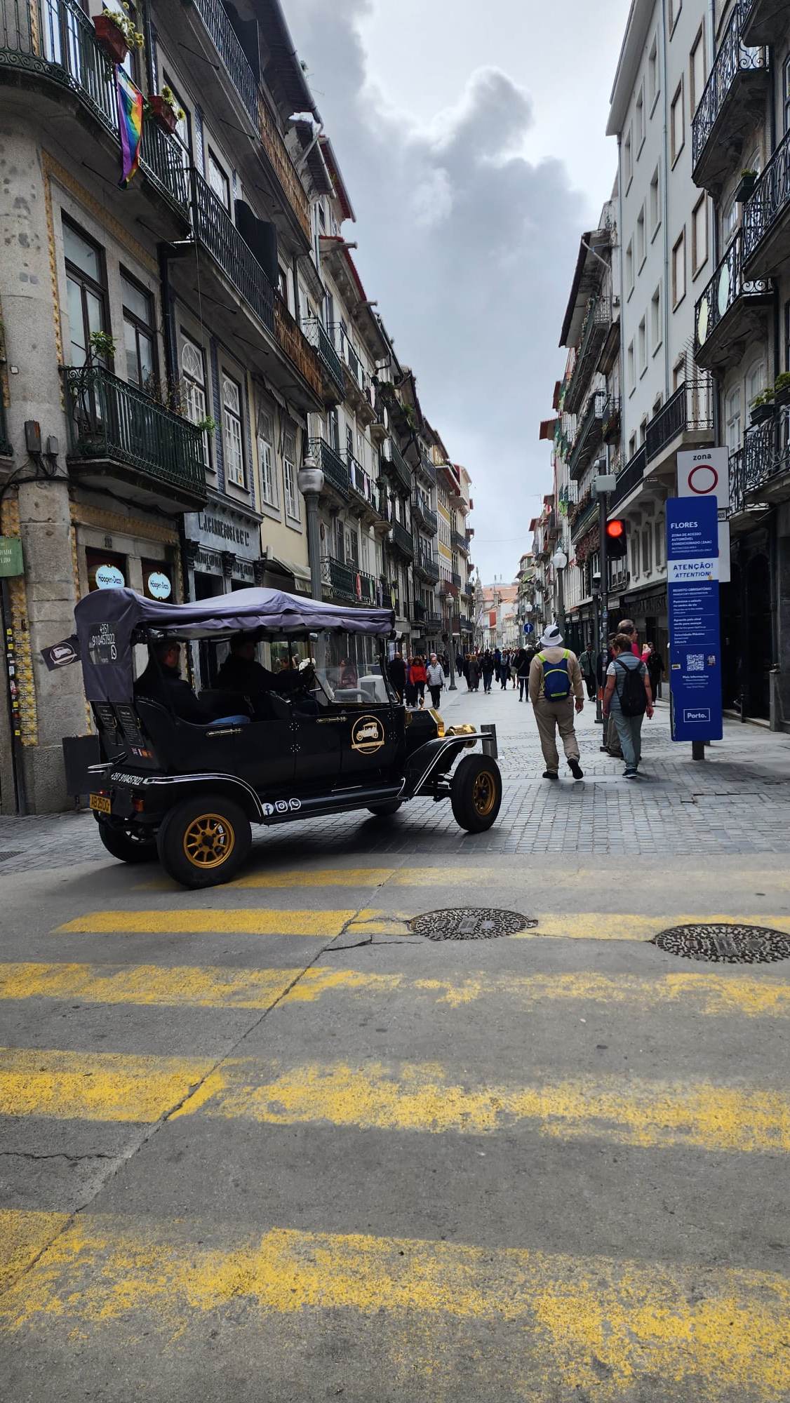 A vintage-style black car with a canopy drives down a cobblestone street lined with historic buildings featuring wrought-iron balconies. A rainbow flag hangs from one balcony, and pedestrians stroll along the street under a cloudy sky.