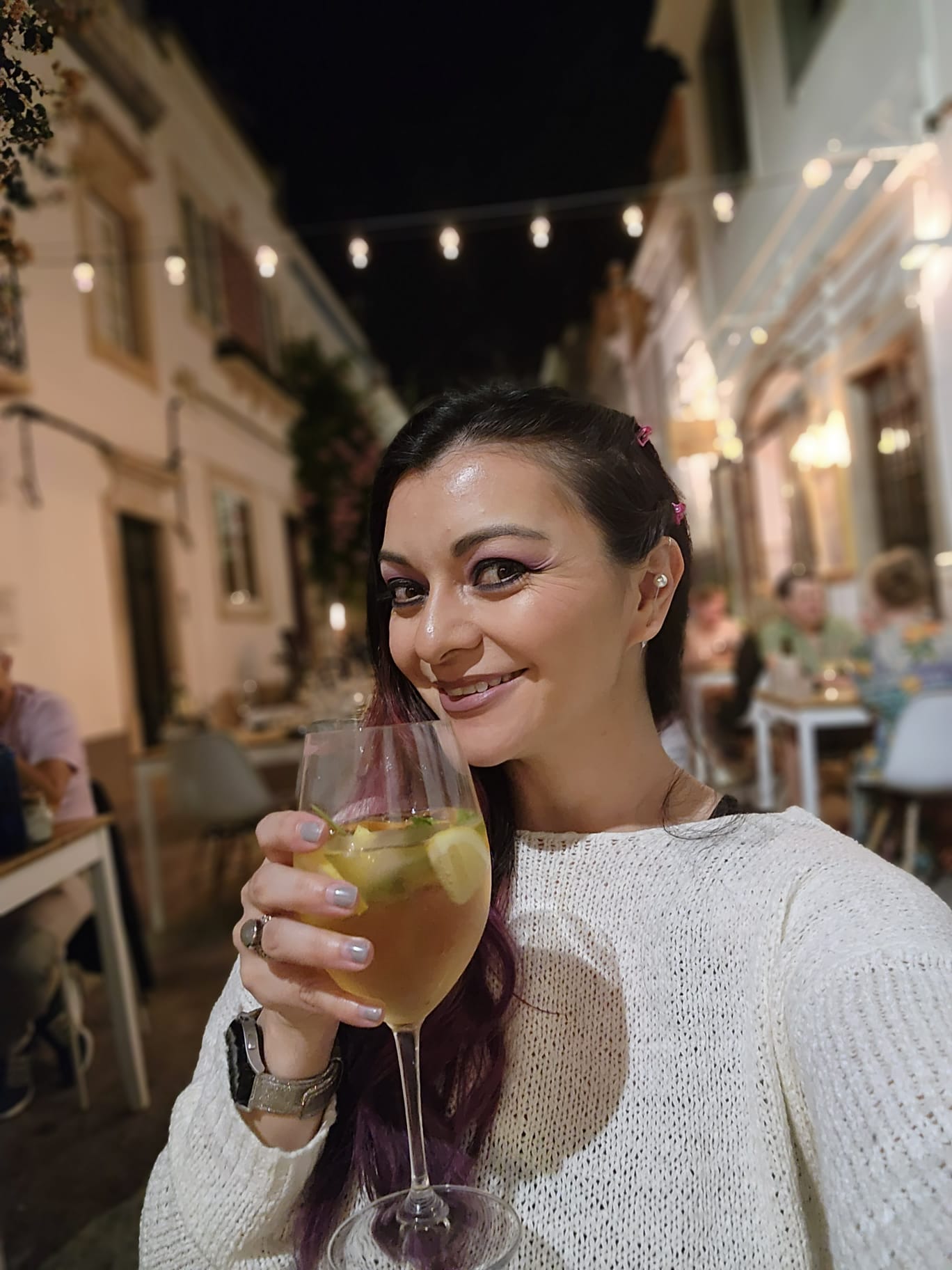 A woman in a white sweater smiles while holding a glass of white wine with lemon slices, seated at an outdoor café at night. The background features a charming street with string lights and diners at tables.