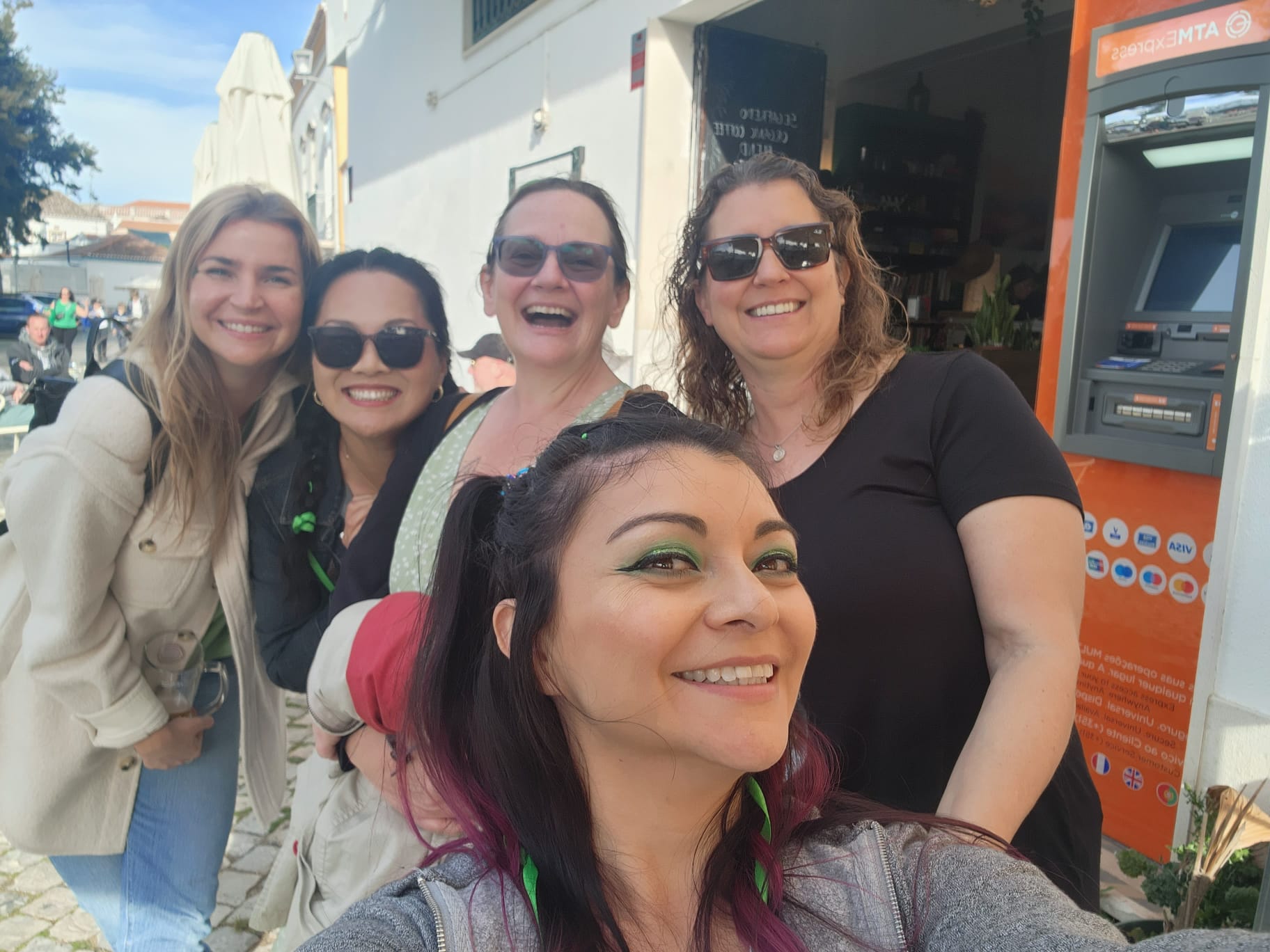 A group of five smiling women take a selfie outdoors near an ATM, with a sunny street and café in the background. They wear casual clothing and sunglasses, enjoying a cheerful moment together.