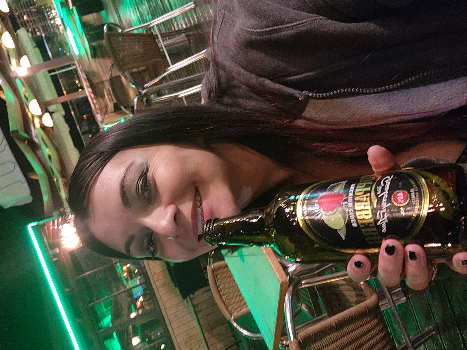 A woman is smiling while holding a bottle of cider with a visible label, seated in an outdoor area with wicker chairs and tables. The setting is warmly lit with green neon accents, creating a lively atmosphere.