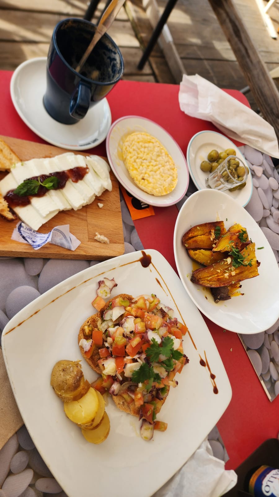 A colorful spread of food on a table includes a plate of bruschetta topped with diced tomatoes and herbs, accompanied by sliced potatoes. Nearby, there are roasted squash pieces garnished with parsley, a small jar of green olives, and a wooden board with sliced cheese and a dollop of jam. A dark blue mug sits on a saucer beside a plate of corn on the cob.