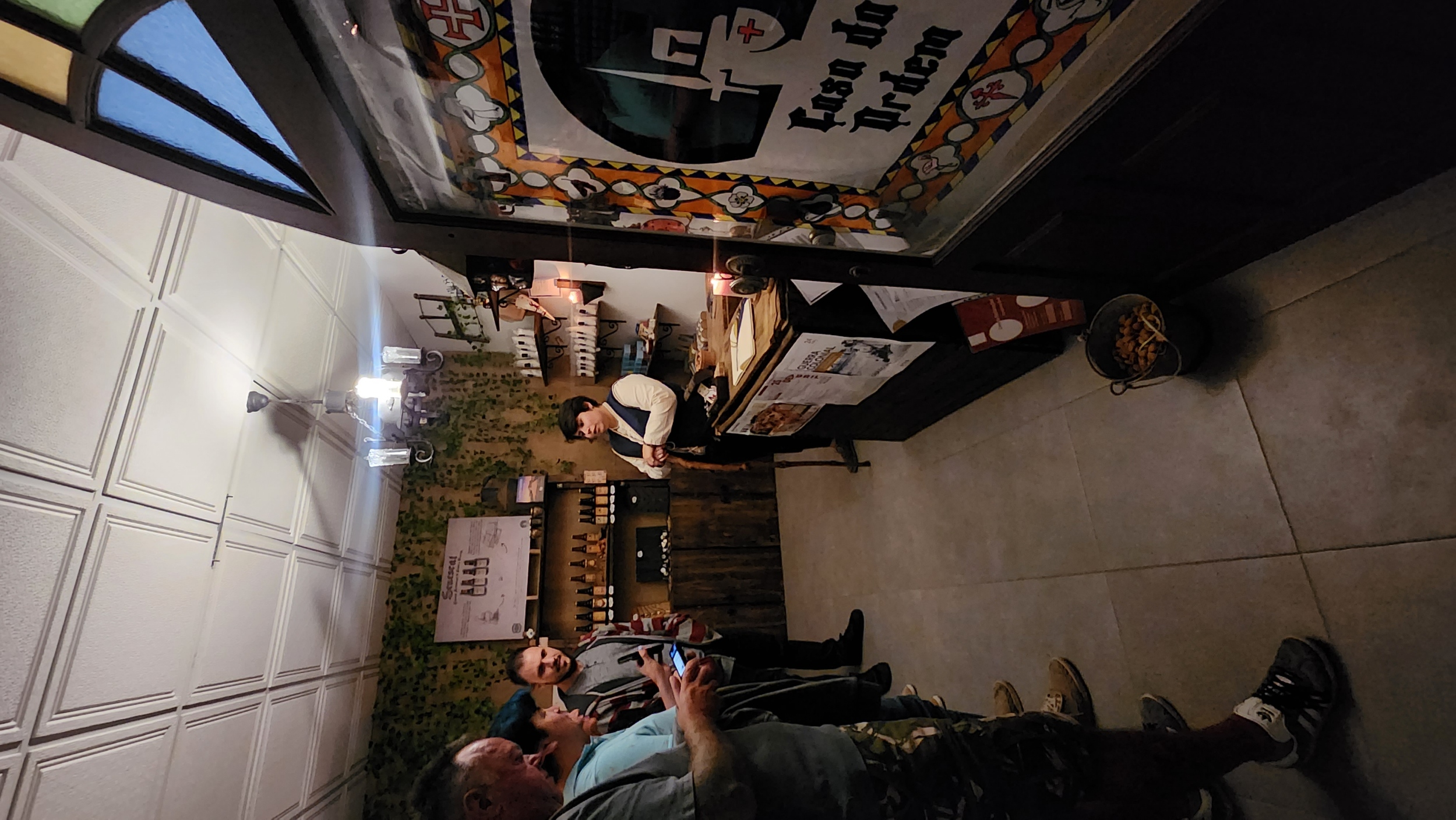 A small shop interior with a person standing behind a counter, talking to four people. The walls are decorated with bottles and greenery, and a sign reads "Casa da Prén." A chandelier hangs from the ceiling, and a bucket with snacks is on the floor near the entrance.