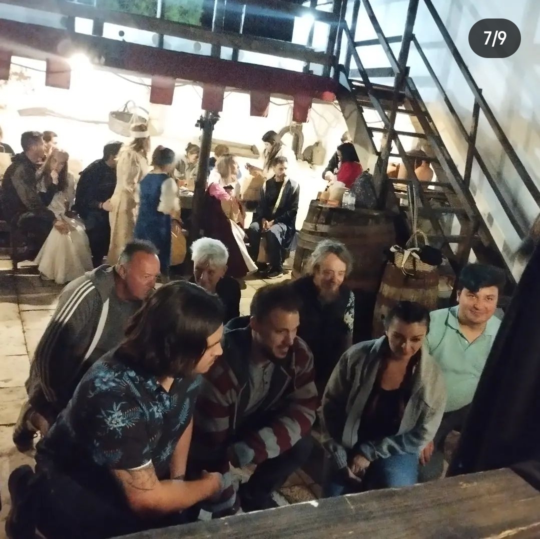A group of people are gathered indoors, some sitting and others kneeling on a wooden floor. In the background, individuals in period costumes are engaged in conversation near a staircase and barrels, creating a lively, historical atmosphere.