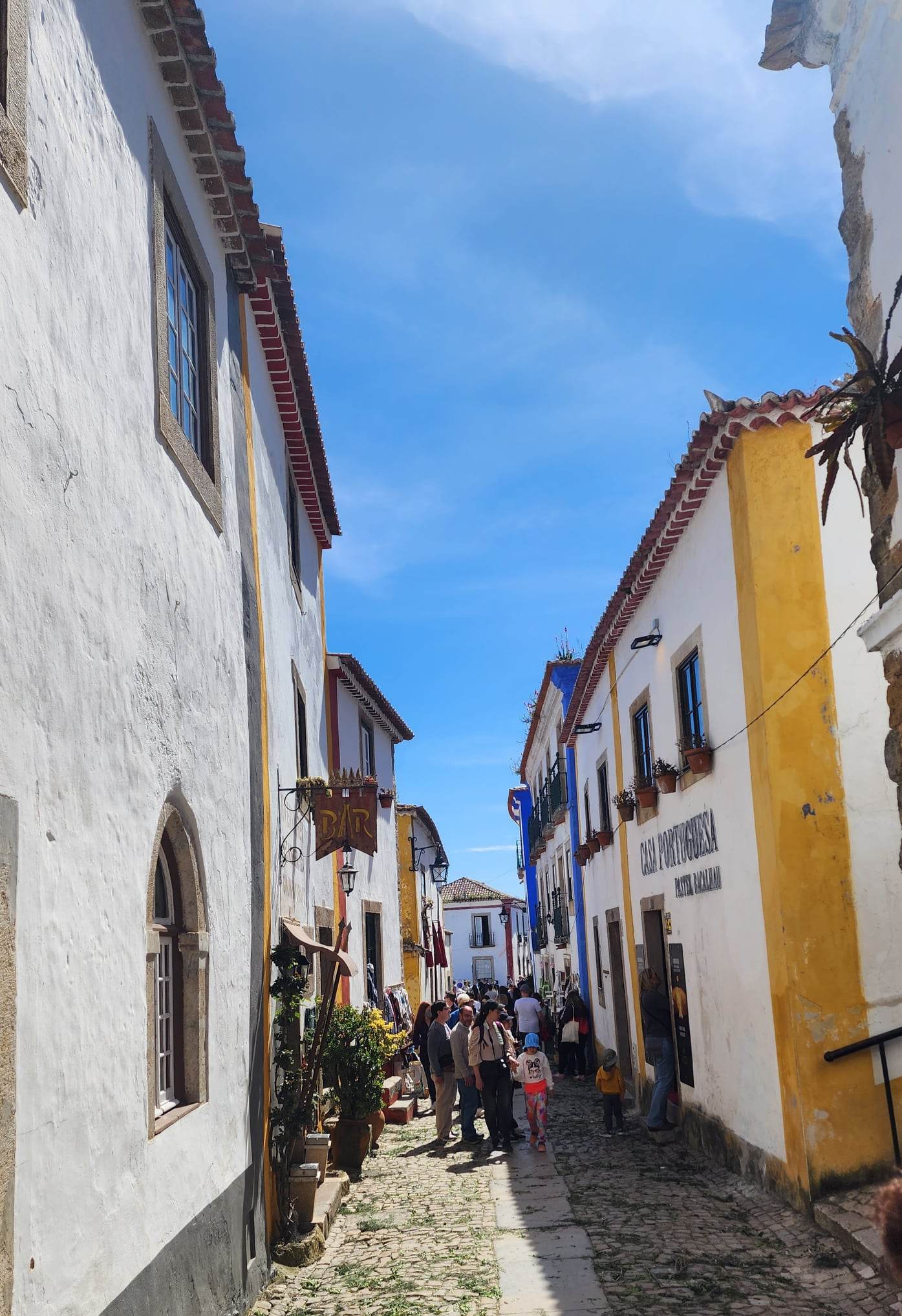 A charming cobblestone street lined with whitewashed buildings featuring yellow accents, bustling with people under a clear blue sky. Signs for a bar and a shop named "Casa Portuguesa" are visible, adding to the quaint, lively atmosphere.