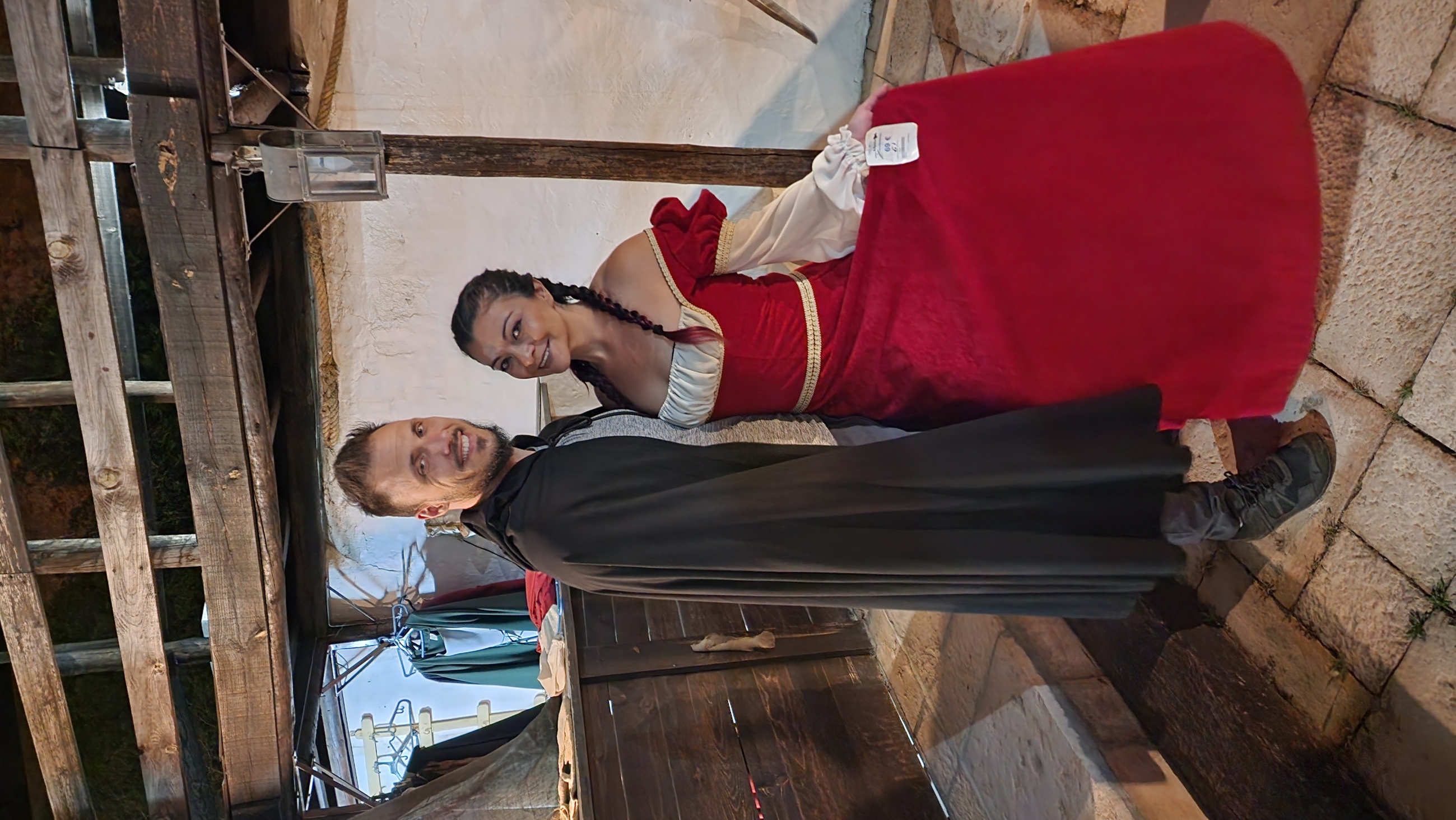A man and woman in period costumes pose together; the man wears a black cloak, while the woman dons a vibrant red and white dress with braided hair. They stand in a rustic setting with wooden beams and stone flooring.