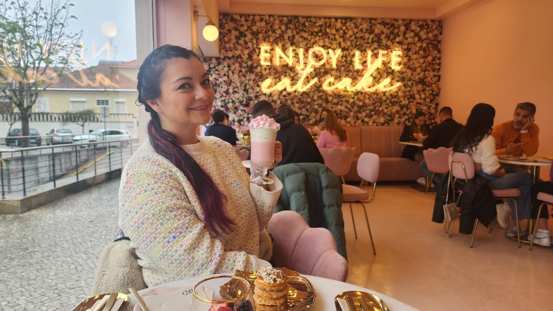 A woman smiles while holding a pink beverage topped with marshmallows, sitting in a cozy café with floral walls and a neon sign reading "Enjoy Life, Eat Cake." The table in front of her features a stack of cookies and a small bowl of berries.
