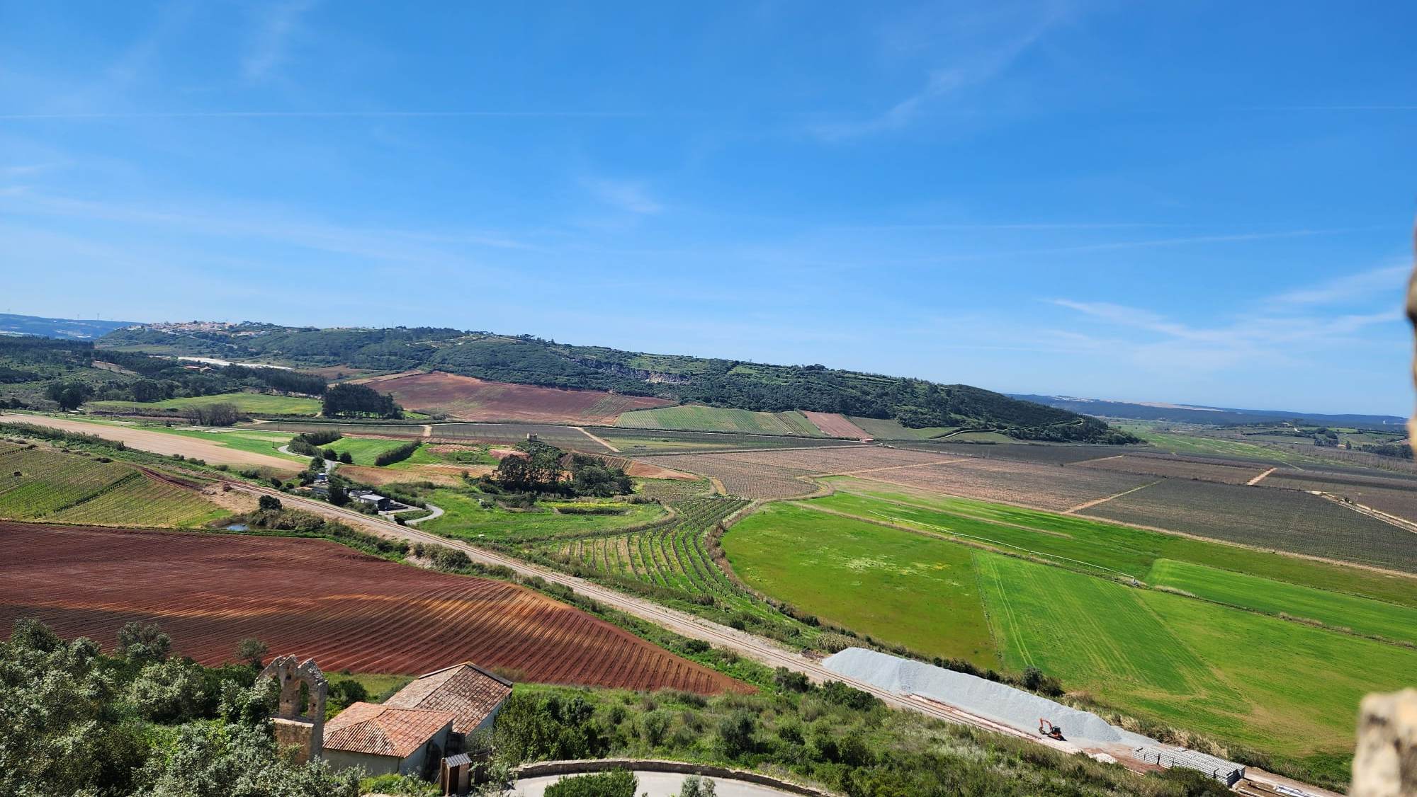 A scenic view of expansive farmland featuring various shades of green and brown fields, gently rolling hills, and a few scattered trees under a clear blue sky. A small building with a red-tiled roof is nestled among the greenery in the foreground.