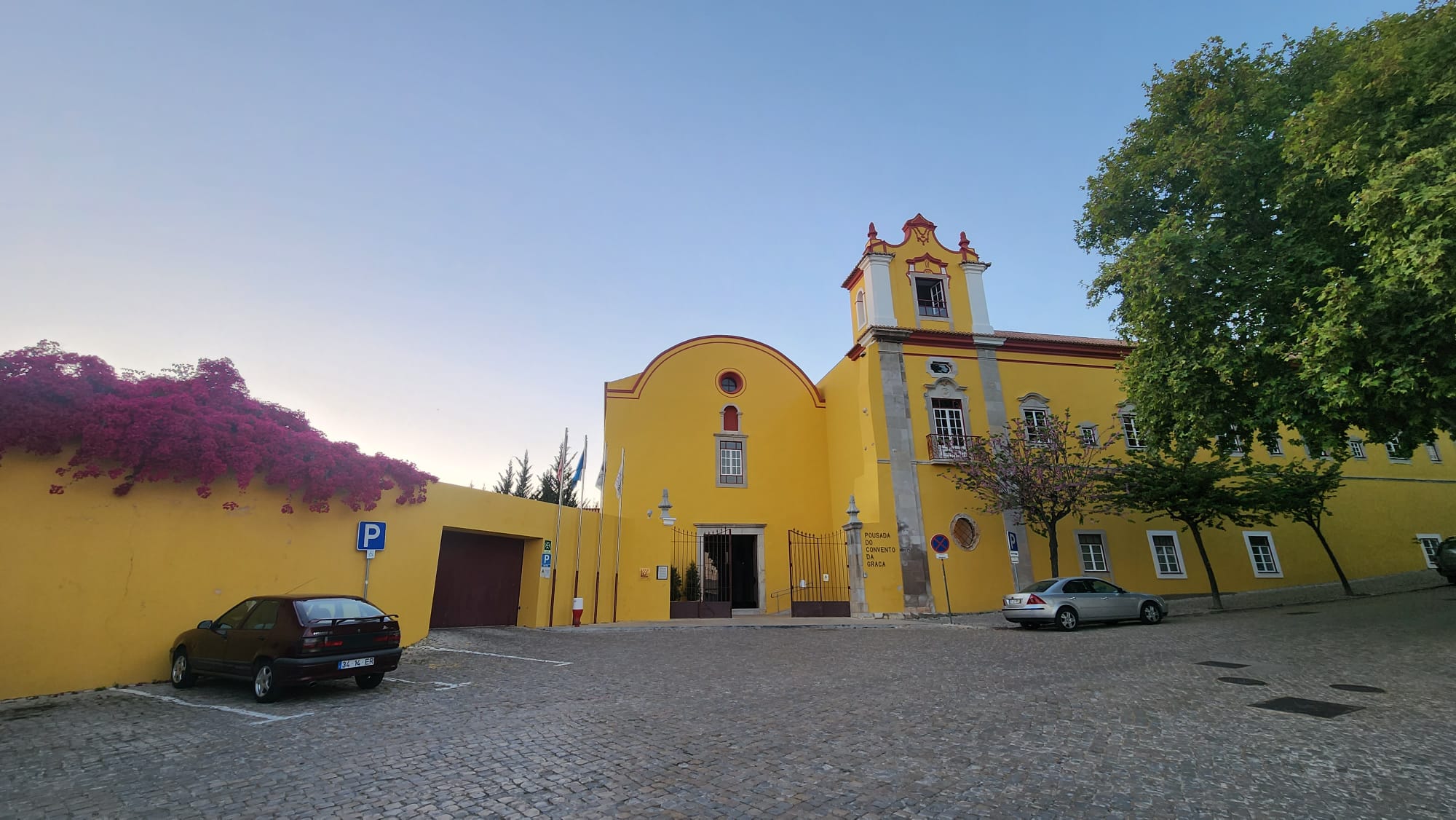 A vibrant yellow building with red accents stands under a clear blue sky, featuring a bell tower and arched entrance. A lush tree and pink flowers adorn the scene, while two parked cars sit on the cobblestone street.