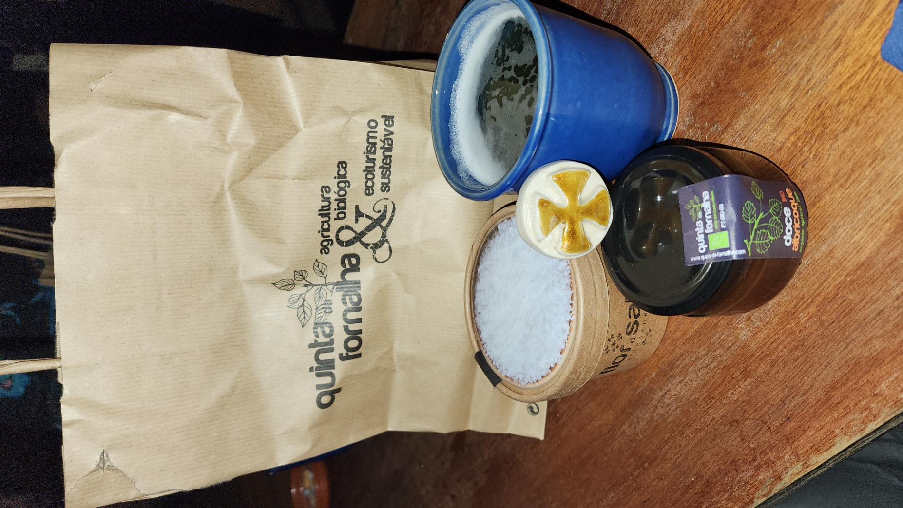 A paper bag labeled "Quinta da Fornalha" stands behind a wooden table with a blue cup, a container of salt, and a jar of organic apricot jam. The scene suggests a focus on sustainable and organic products.