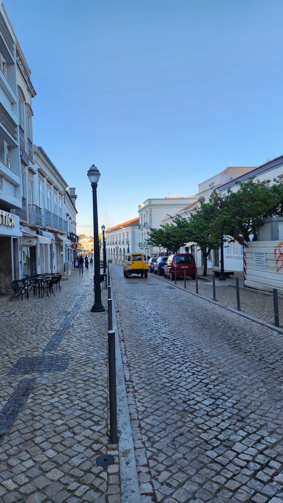 A cobblestone street lined with classic buildings and trees, featuring a yellow vintage car parked among other vehicles. A few people walk along the sidewalk, and the sky is clear and blue, enhancing the quaint atmosphere.
