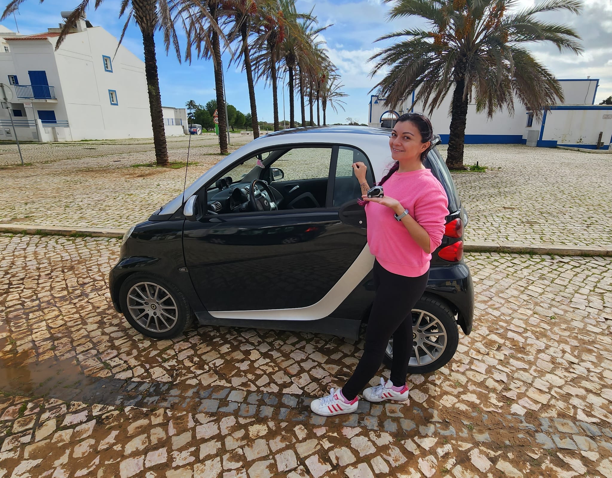 A woman in a pink sweater and black leggings stands smiling beside a small black and silver car, holding the keys. She's in a cobblestone area lined with palm trees, with white buildings in the background.