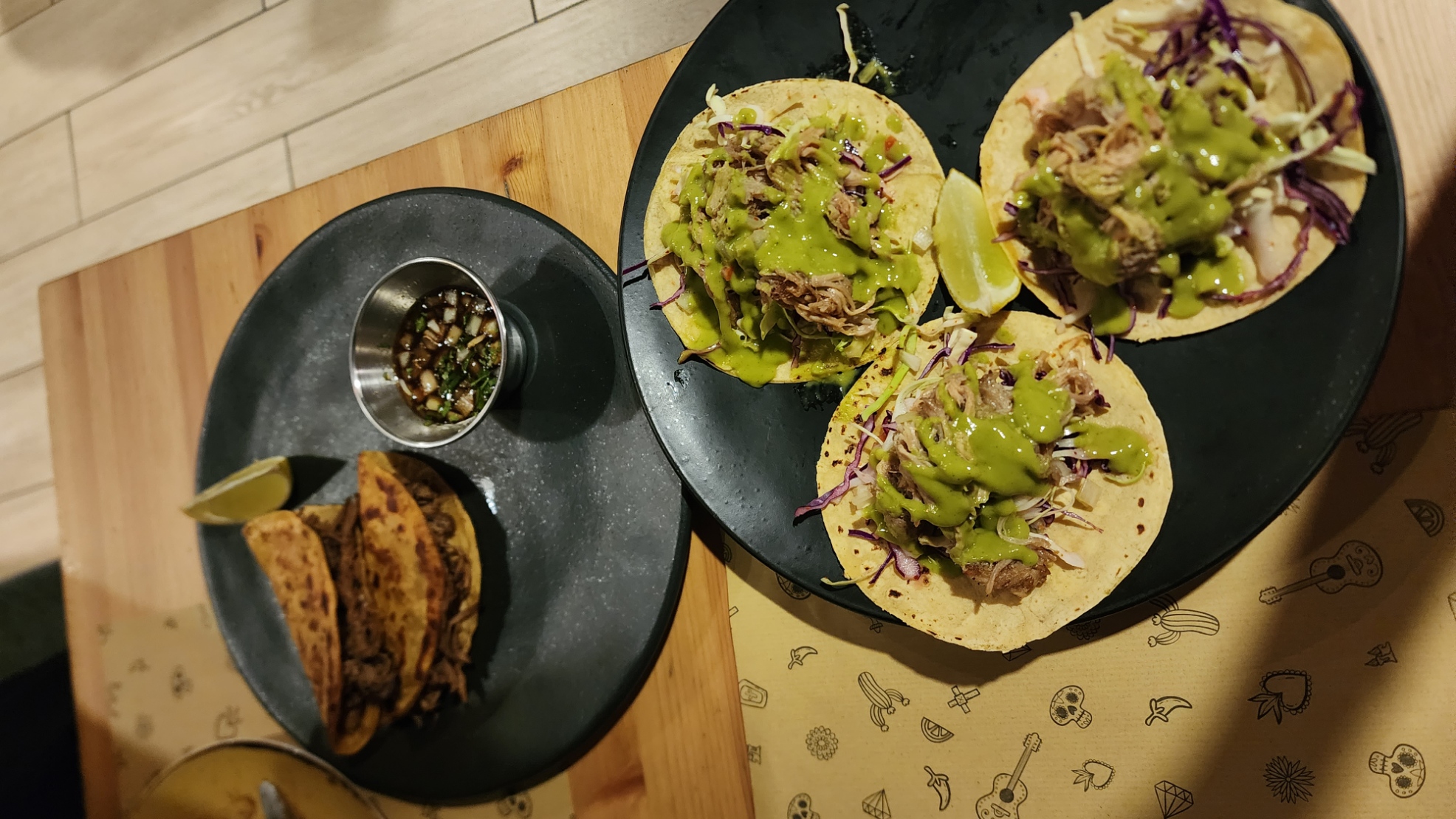 Two plates of delicious tacos are on a wooden table. One plate has crispy tacos with a lime wedge and a small metal cup of sauce. The other plate features soft tacos topped with shredded meat, vibrant purple cabbage, and a drizzle of green sauce, accompanied by lime wedges.