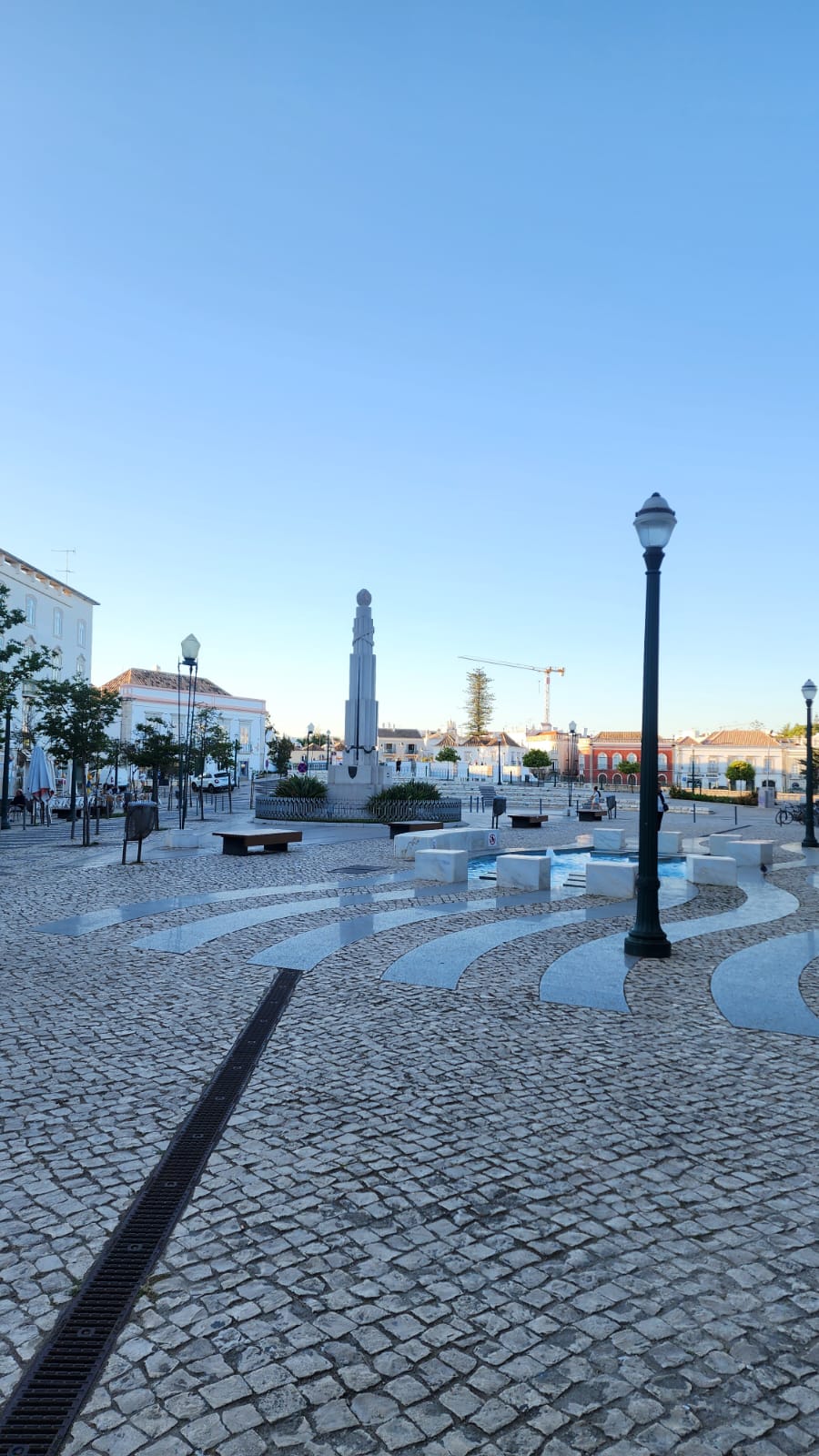 A tranquil town square features a tall, slender monument surrounded by benches and trees. The cobblestone ground is patterned with curved stone paths, and several vintage-style street lamps are visible. The sky is clear and blue, adding to the serene atmosphere.