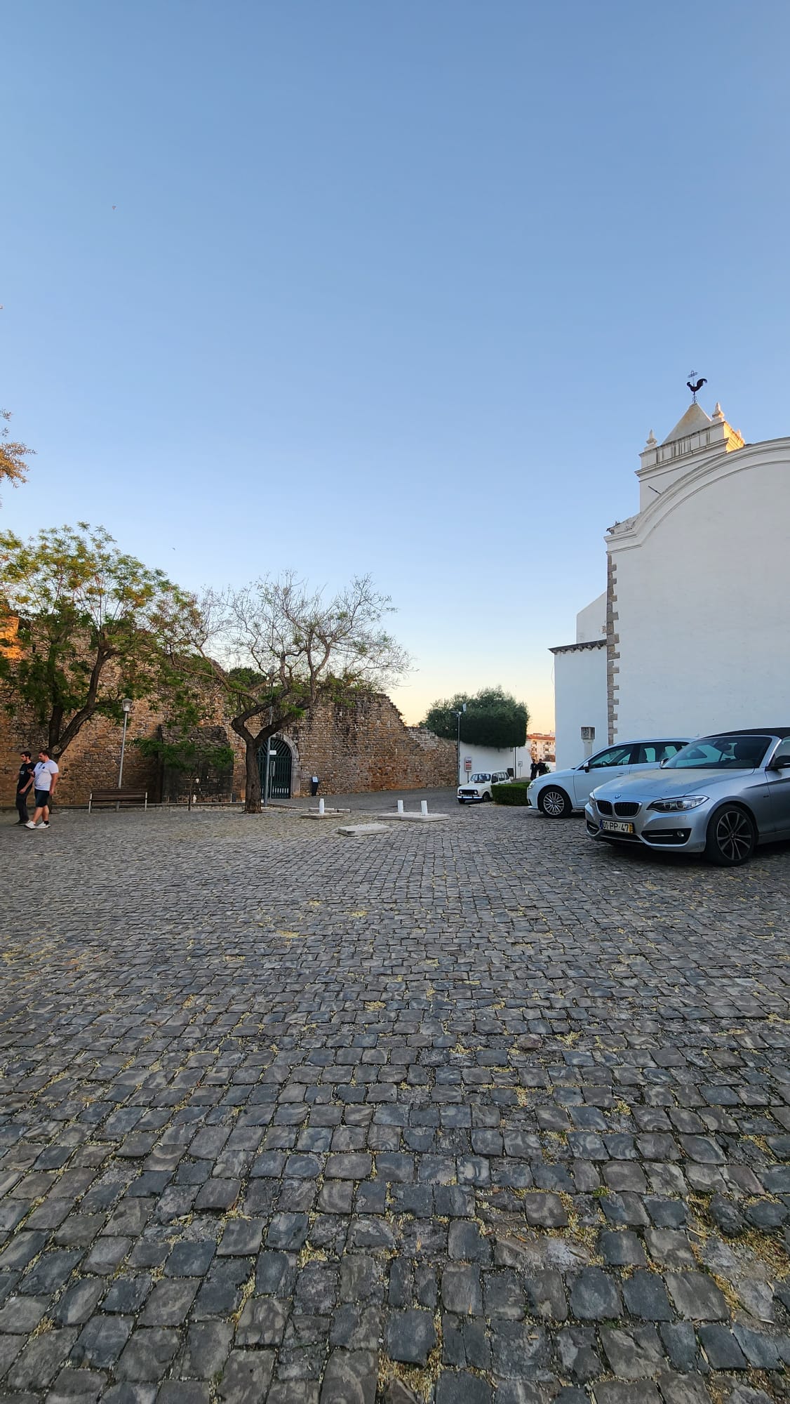 A cobblestone courtyard with a clear blue sky, featuring a white building with a decorative rooftop on the right. Two cars are parked nearby, and two people are walking beside a tree and an ancient stone wall in the background.