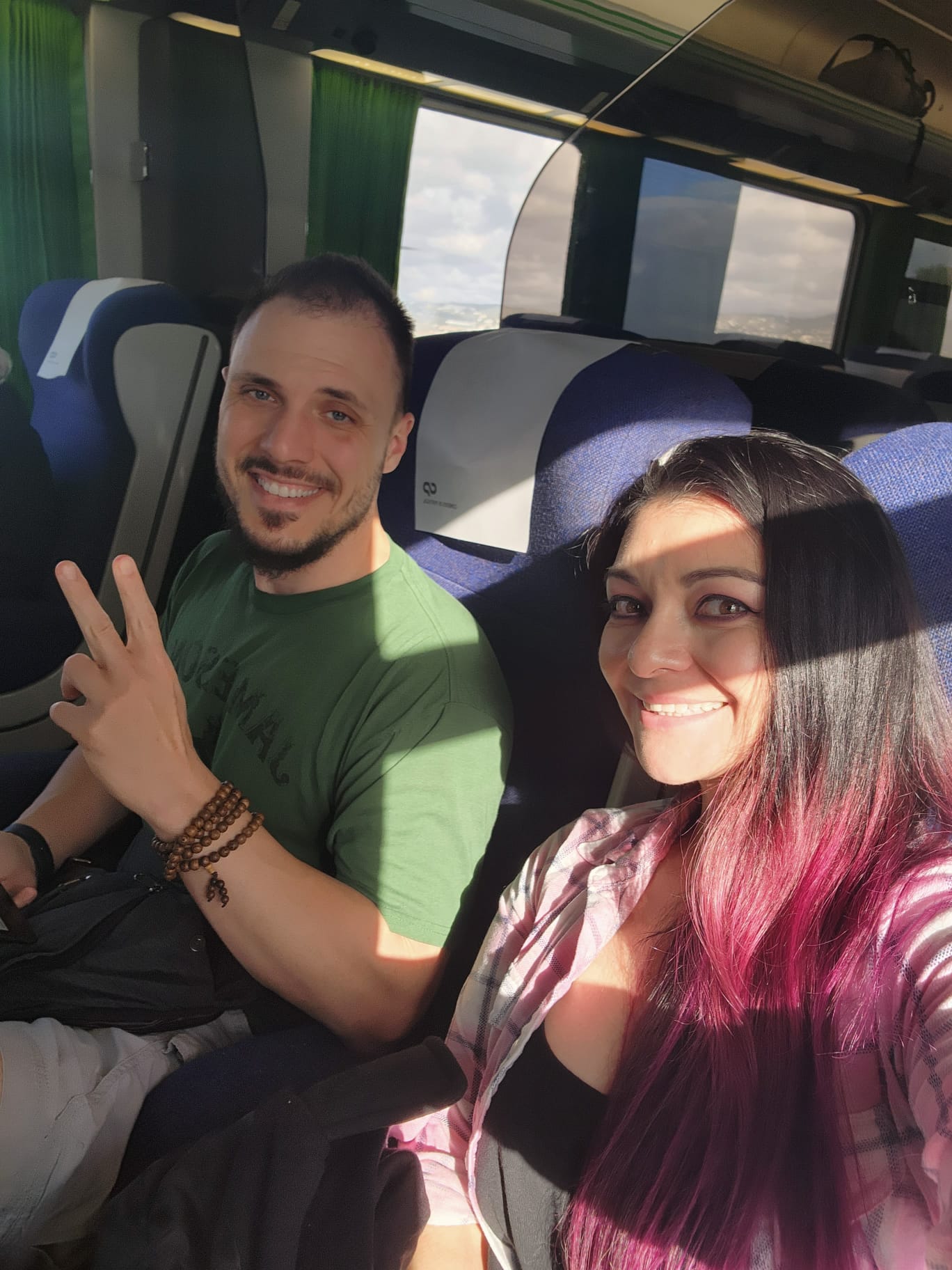 A man and a woman are sitting together on a train, smiling at the camera. The man is wearing a green shirt and making a peace sign, while the woman has long pink-tinted hair and is wearing a plaid shirt. Sunlight streams through the window, casting a warm glow inside the carriage.