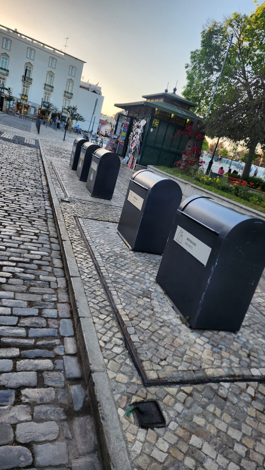 A row of black recycling bins is lined up on a cobblestone street in a quaint urban setting, with a historic building and a kiosk in the background. A tree with lush green foliage stands nearby, and people are visible in the distance under a clear sky.