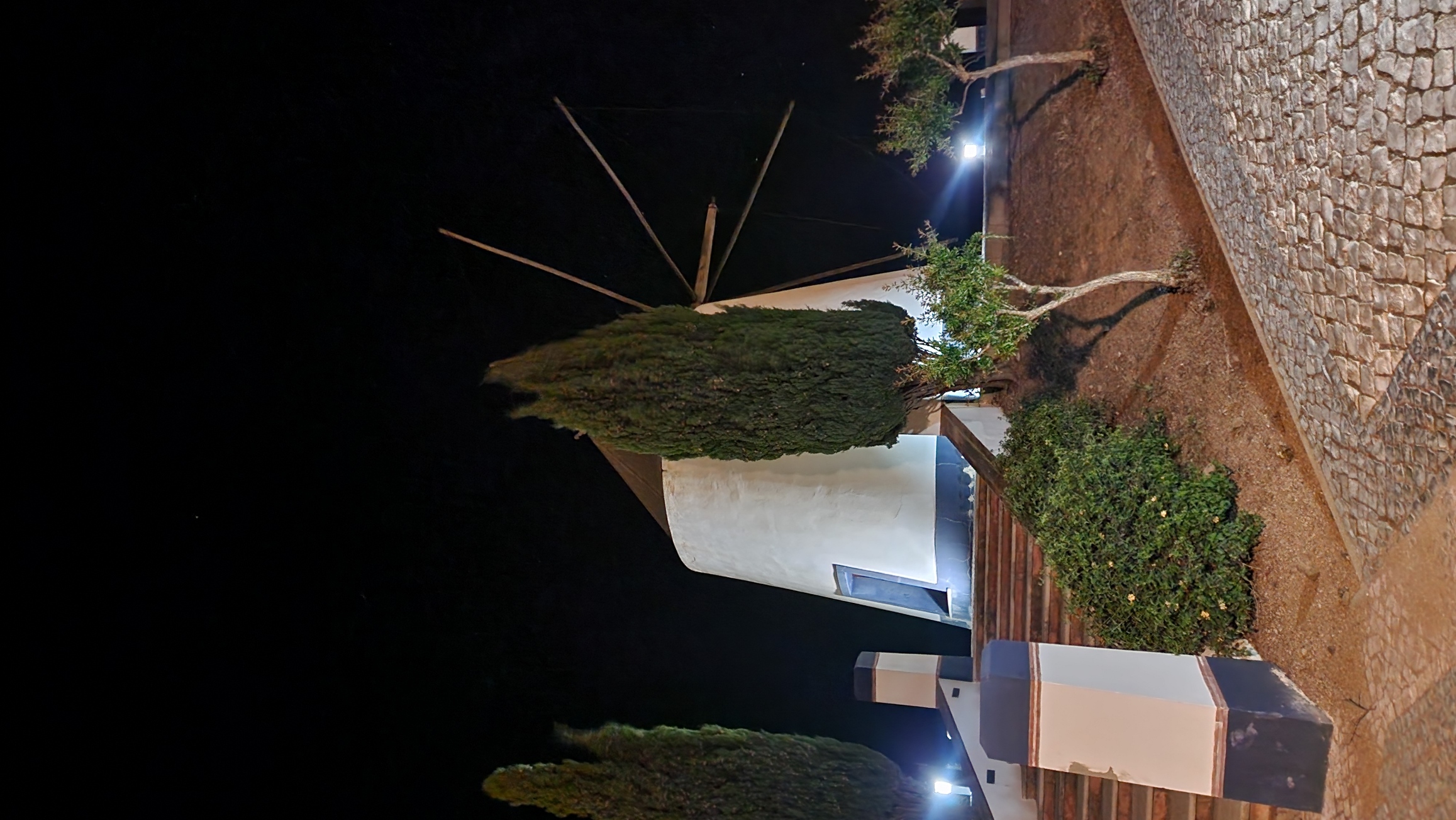 A traditional windmill stands illuminated at night, with its white cylindrical structure and wooden blades visible. Tall cypress trees flank the windmill, and a cobblestone path leads up to it, creating a serene and picturesque scene.