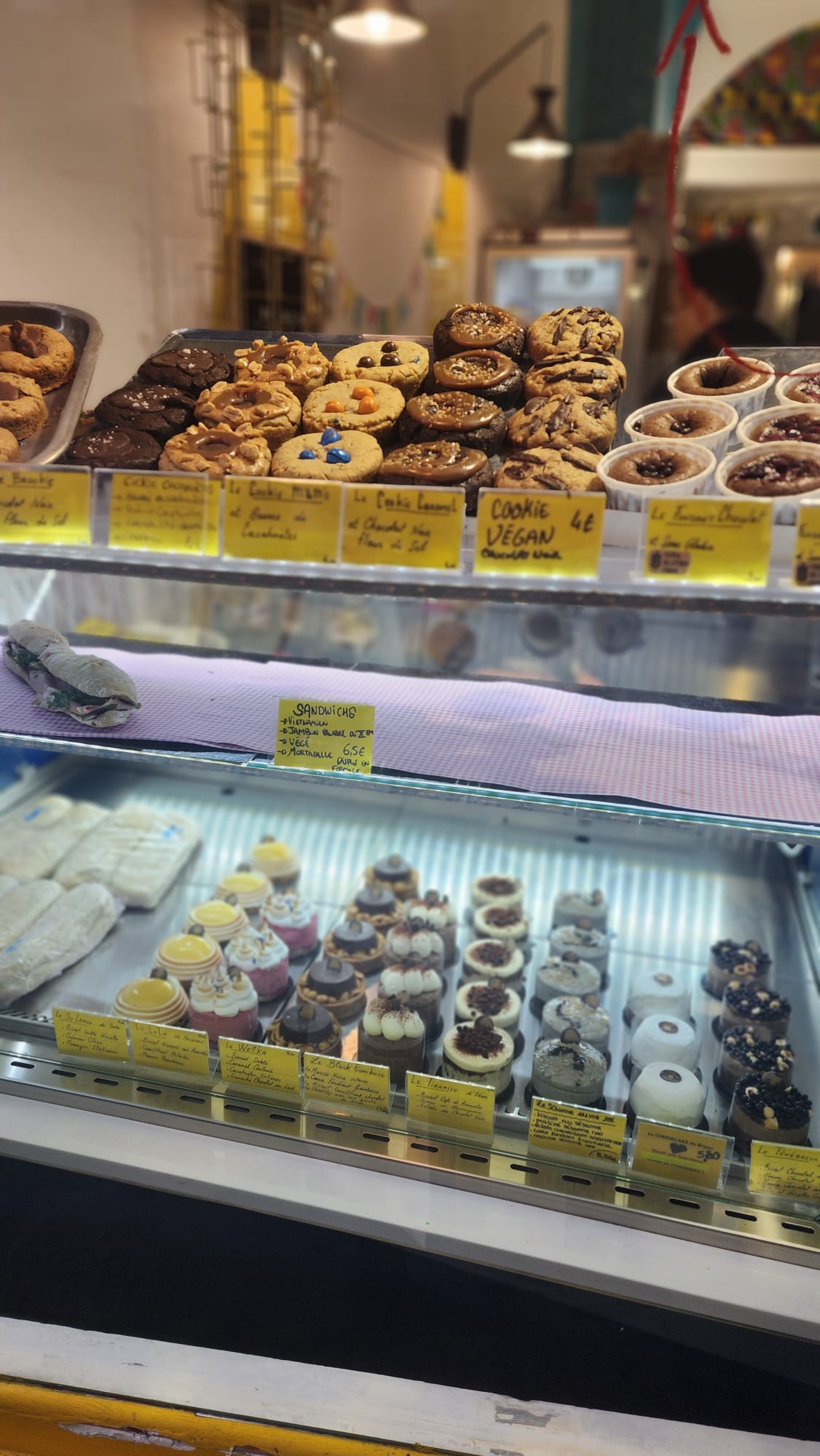 A display case filled with assorted baked goods, including a variety of cookies on the top shelf, some labeled as vegan. Below, there are colorful cupcakes and pastries, each with detailed labels. The setting appears to be a cozy bakery with a warm ambiance.