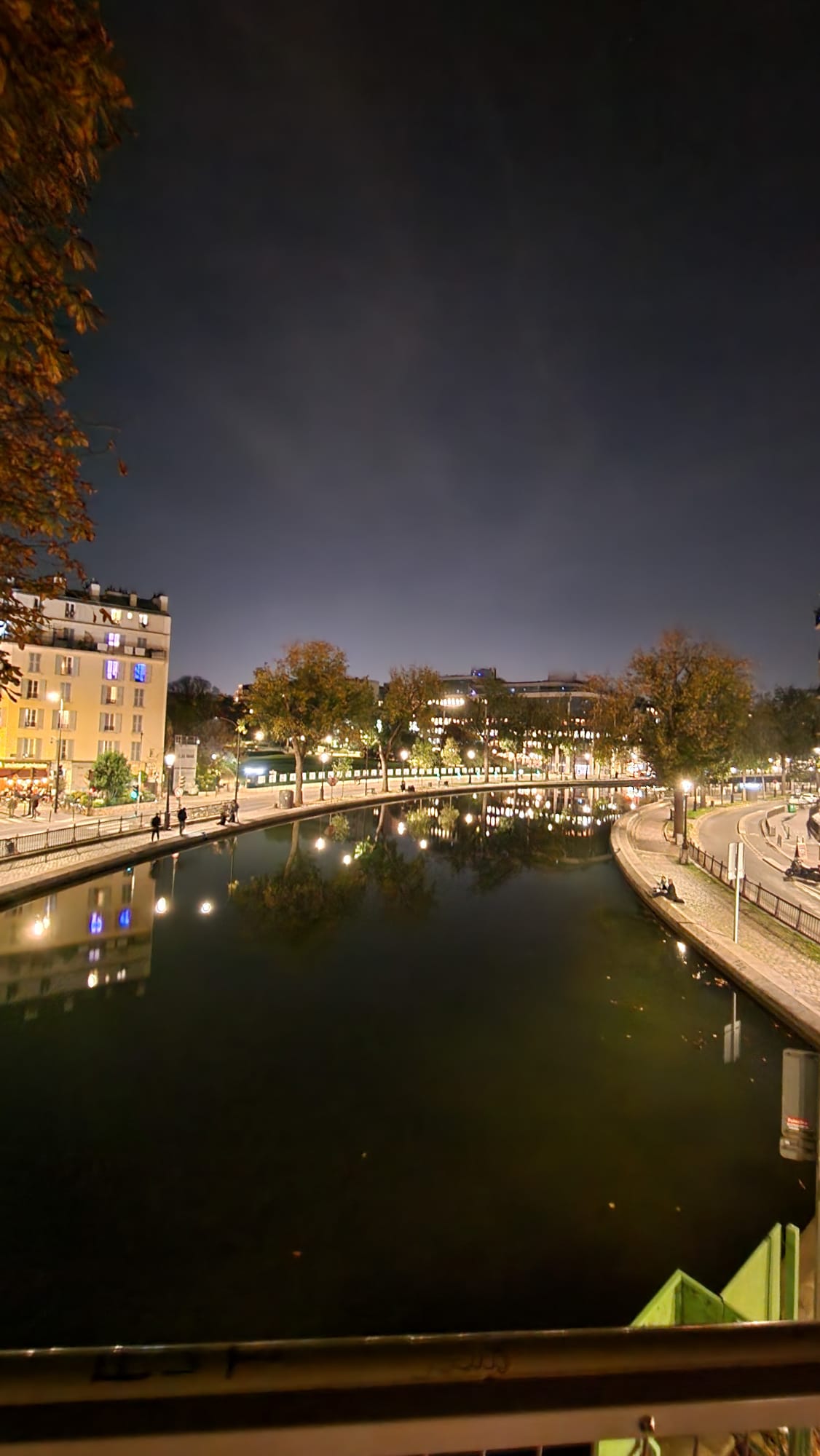 A nighttime view of a serene canal lined with trees and illuminated by streetlights, reflecting in the water. To the left, a row of buildings with glowing windows adds warmth to the scene, while a few people stroll along the cobblestone path.