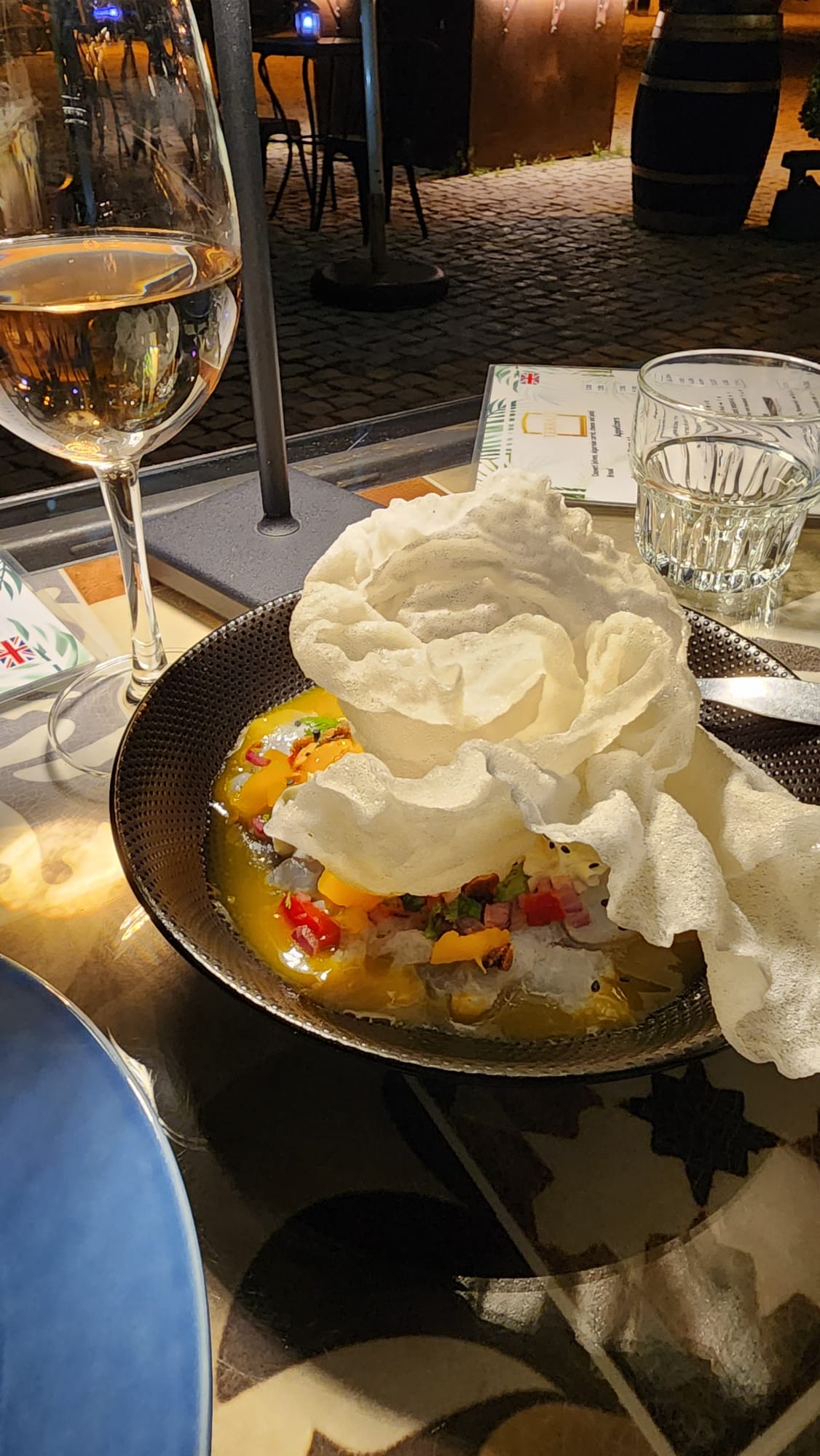 A glass of white wine and a water glass sit on a table beside a dish featuring a vibrant ceviche topped with a large, crispy white cracker. The dish is garnished with colorful diced vegetables, and the setting appears to be an outdoor dining area with cobblestone flooring and warm lighting.