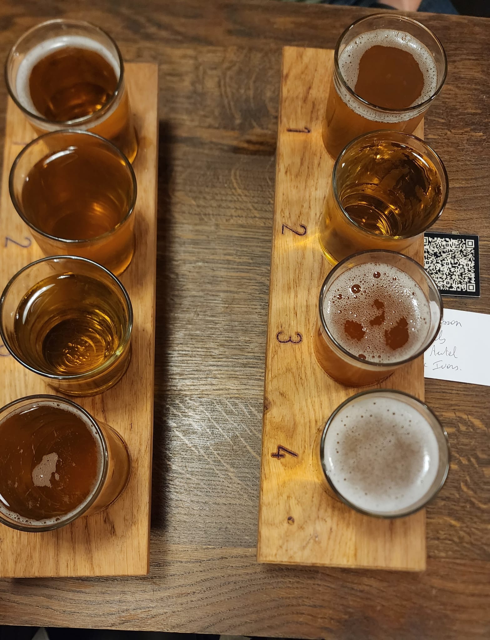 Two wooden trays each hold four glasses of beer, numbered 1 to 4, on a rustic wooden table. The beers vary in color, ranging from light amber to a deeper golden hue, with foamy heads visible on some. A QR code and a small piece of paper are partially visible in the background.