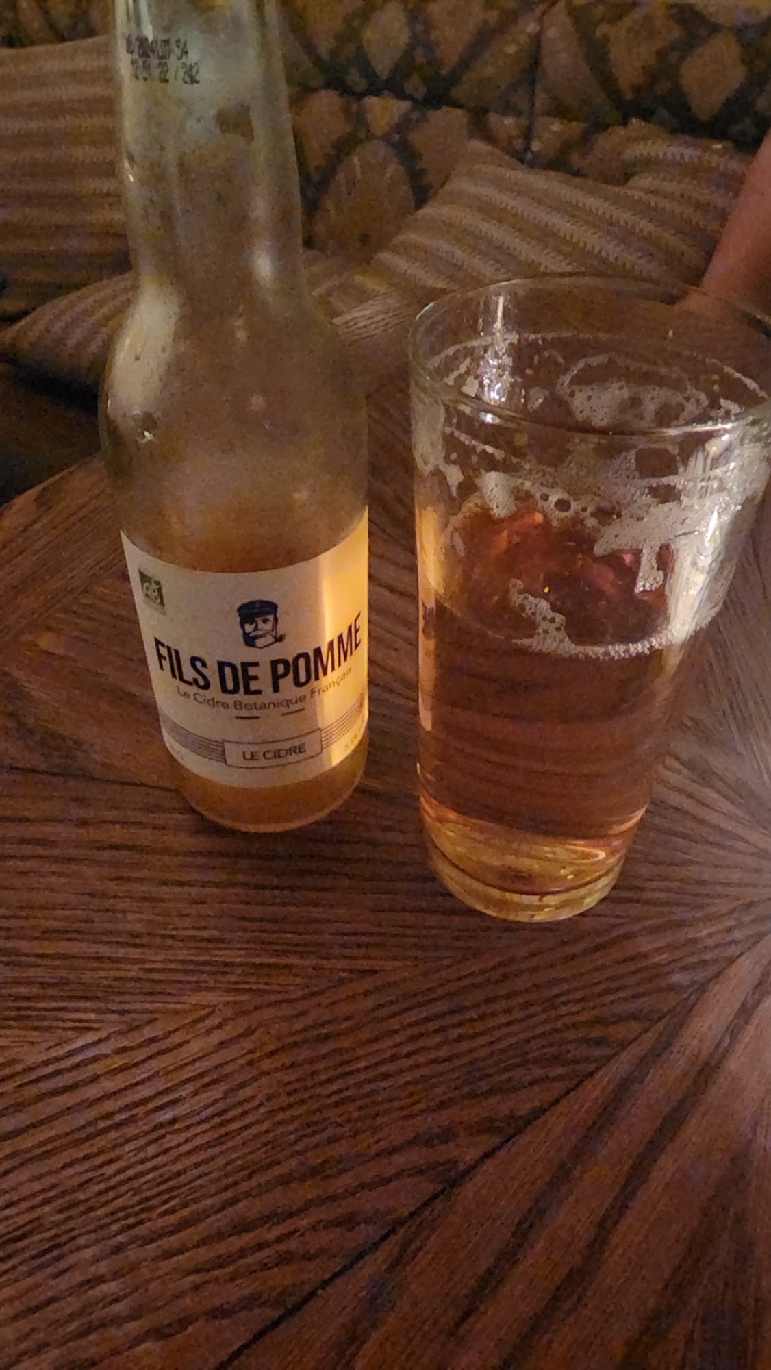 A bottle of "Fils de Pomme" cider sits next to a half-filled glass on a wooden table, with a cozy, patterned couch in the background.