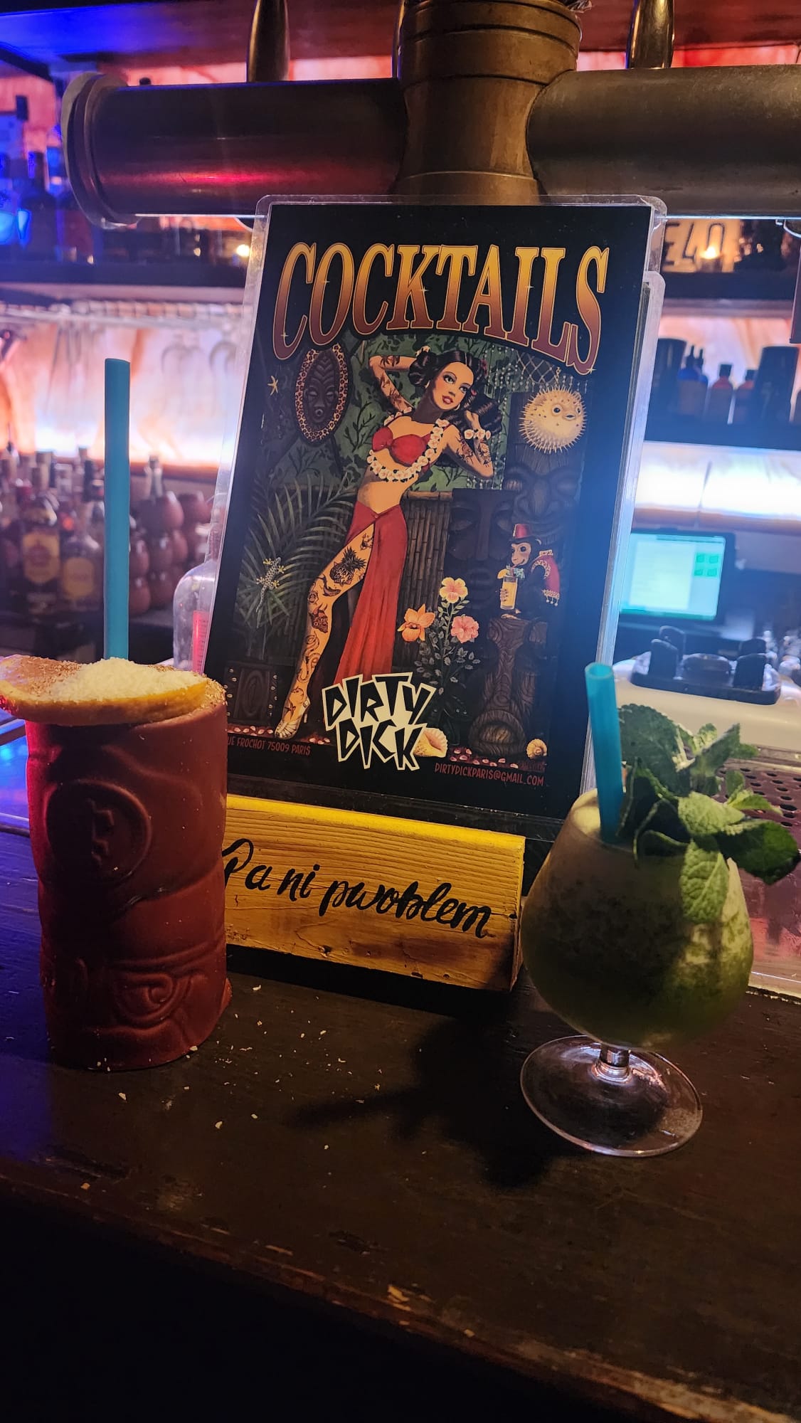 Two cocktails sit on a dimly lit bar; one in a red tiki glass with a blue straw and a citrus garnish, and the other in a rounded glass with mint leaves. A colorful cocktail menu with a vintage illustration is displayed behind them.