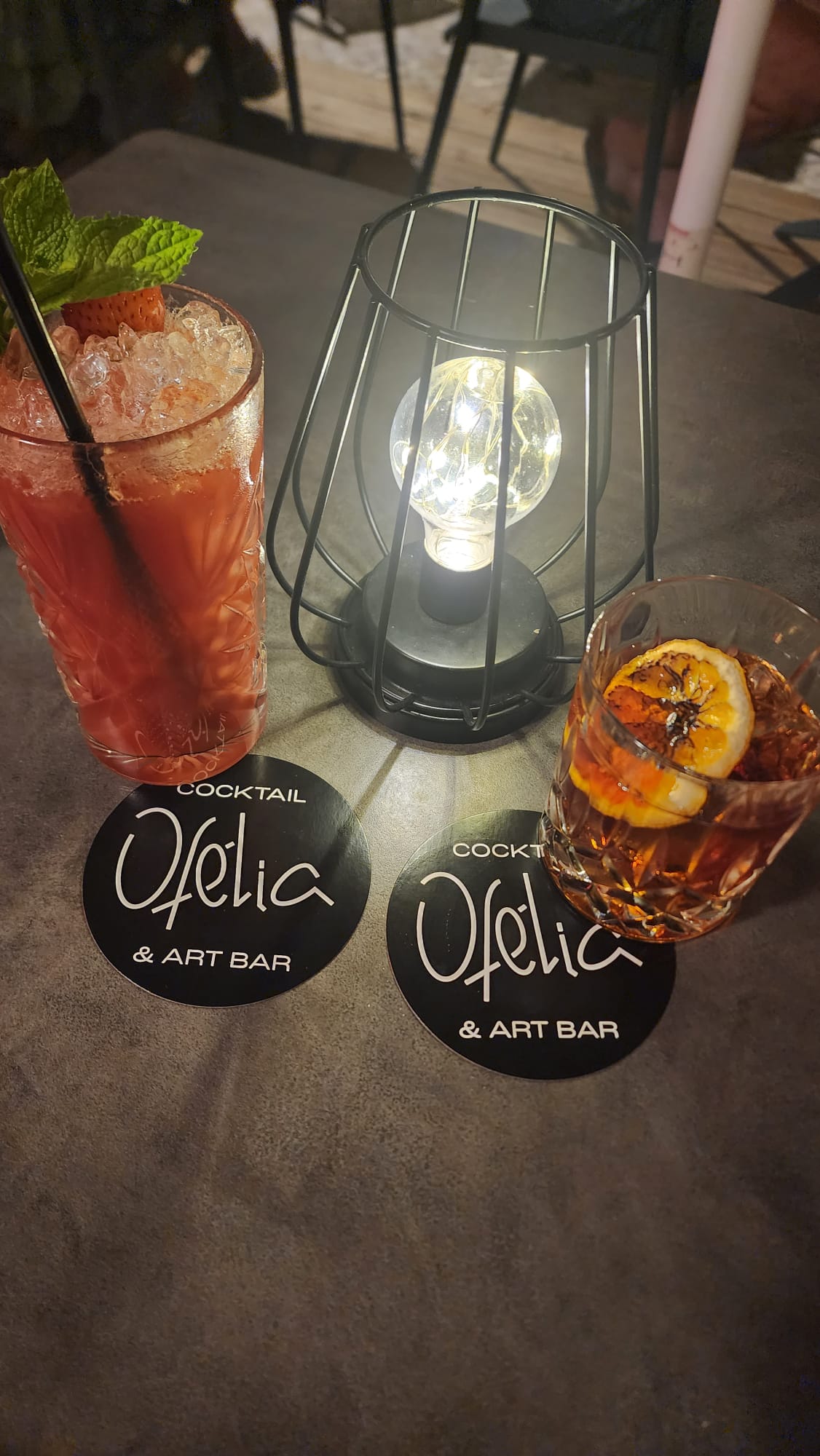 Two cocktails sit on a table at Ofelia Cocktail & Art Bar, one red with mint and a strawberry garnish, the other with a slice of citrus on top. A small decorative lamp adds a cozy ambiance.