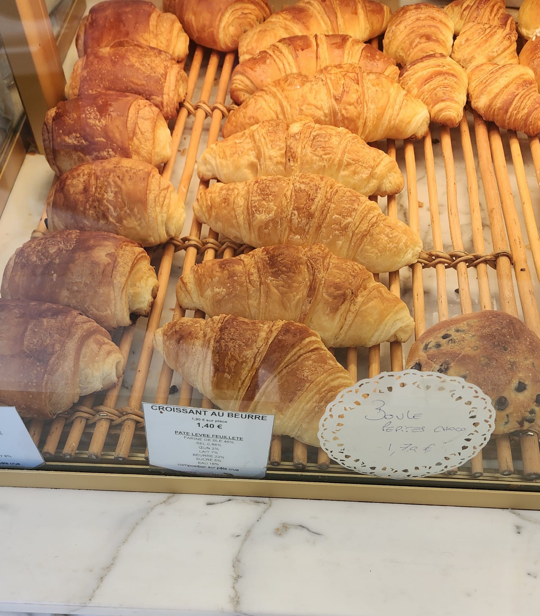 A display case filled with golden, flaky croissants and chocolate pastries on a wooden rack, with price tags indicating "Croissant au Beurre" for 1.40€ and "Boule Pépites Choco" for 1.70€.
