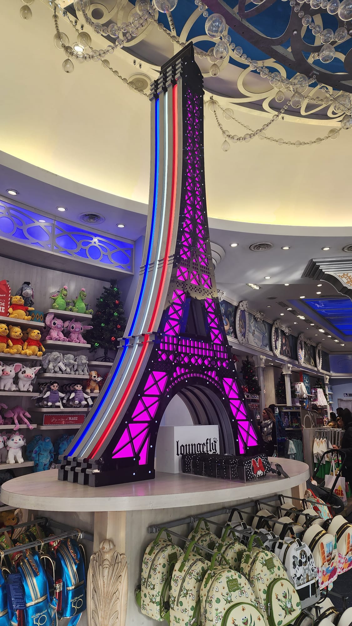 A vibrant Eiffel Tower replica with colorful lights stands in a store, surrounded by shelves filled with plush toys and a variety of themed backpacks. The ceiling is adorned with decorative elements and hanging ornaments, creating a festive atmosphere.