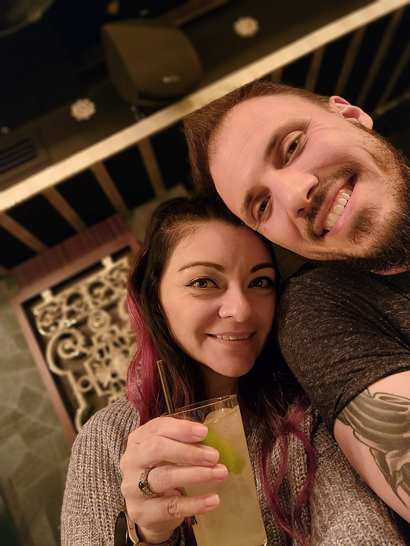 A smiling couple takes a close-up selfie in a warmly lit setting, with the woman holding a drink garnished with a lime slice. She has long hair with a hint of pink, and the man has a beard and a visible tattoo on his arm.