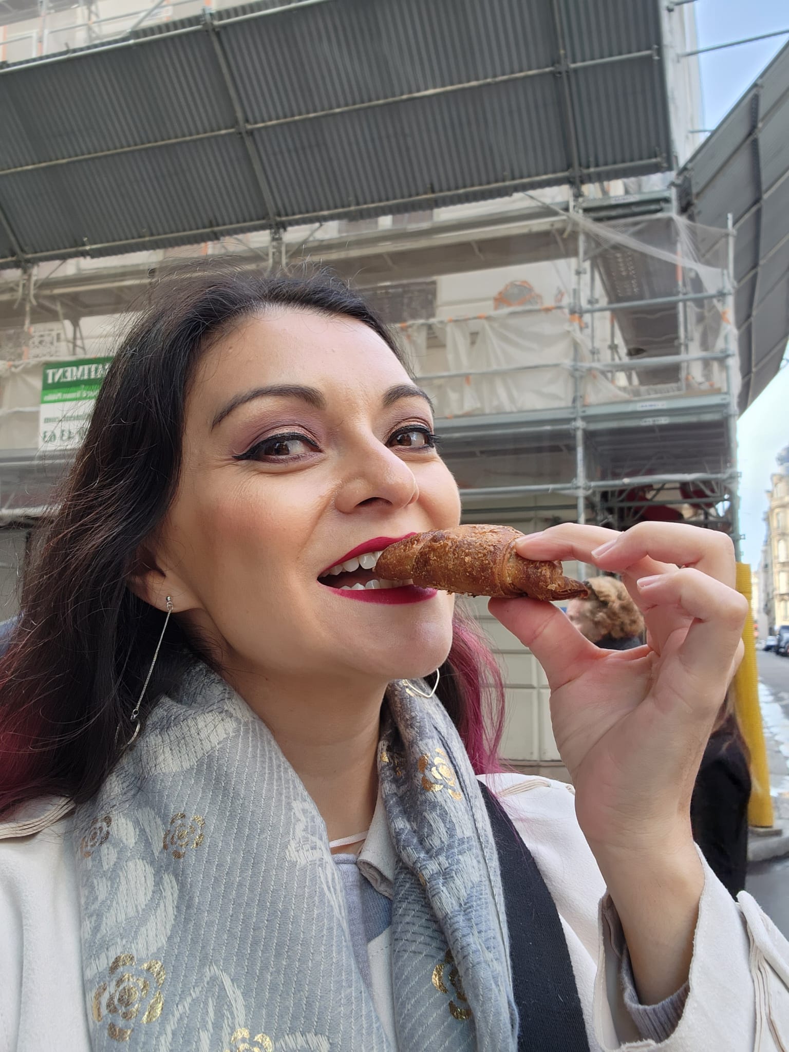A woman with dark hair and red lipstick is about to take a bite of a small pastry while standing on a city street. She is wearing a light-colored coat and a patterned scarf, with a construction site visible in the background.