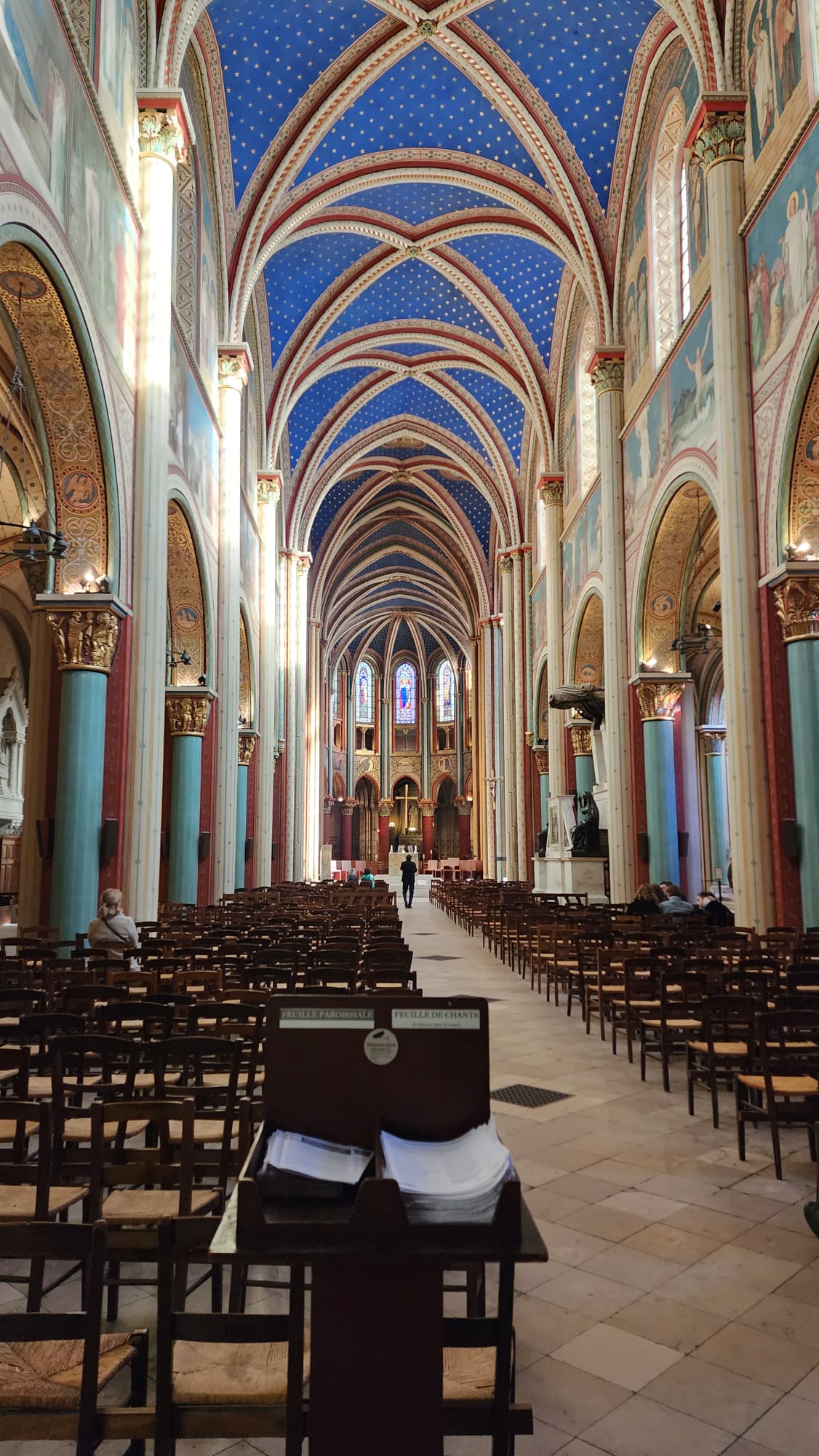 The interior of a grand cathedral features a stunning blue vaulted ceiling adorned with stars, flanked by tall, ornate columns and arches. Rows of wooden chairs line the aisle, leading towards an altar with colorful stained glass windows in the background.